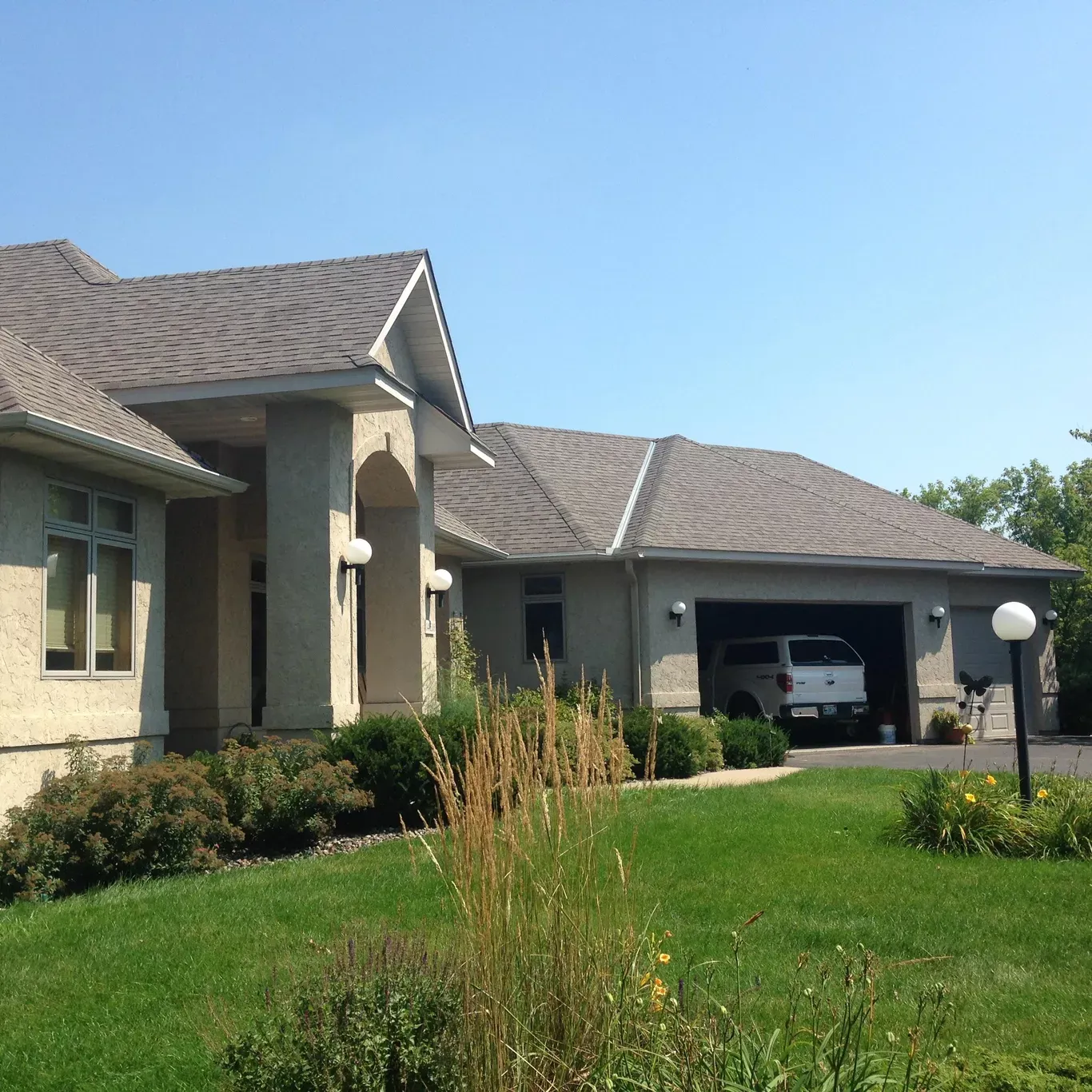 Tan stucco house with gray roof, white truck in open garage, green lawn, blue sky.