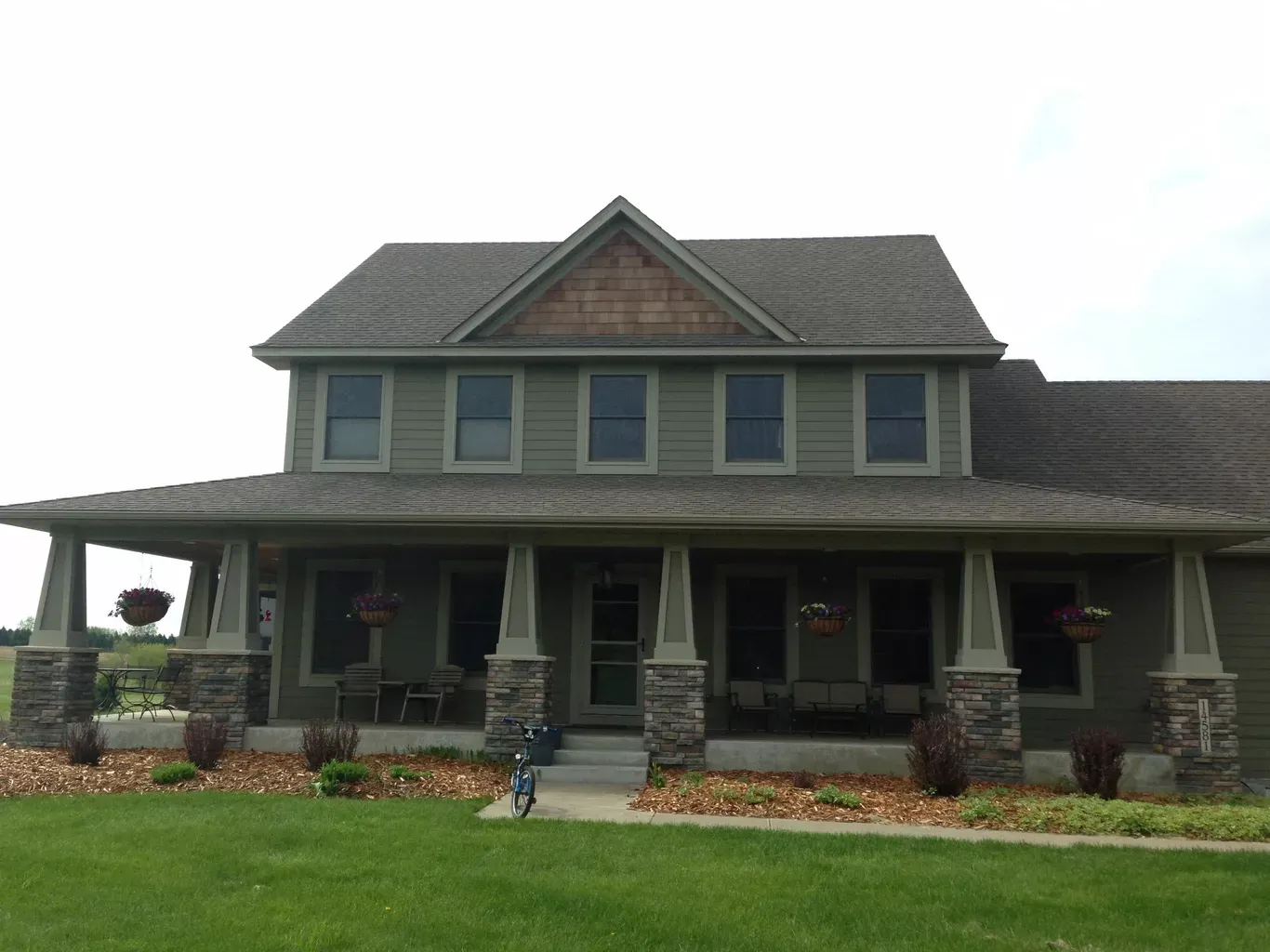 Two-story green house with a wraparound porch, stone columns, and a grassy lawn. Hanging flower baskets decorate the porch.