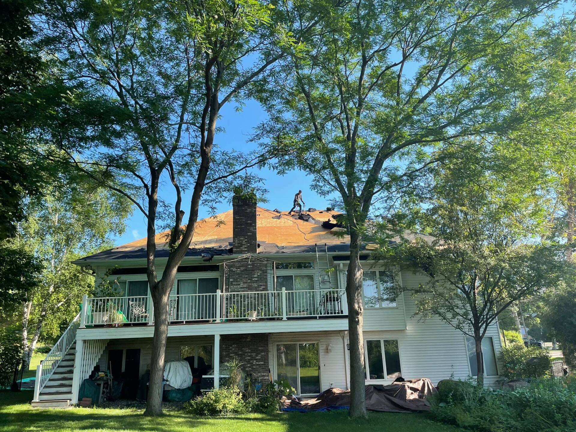 House being worked on; a person is on the roof, with a chimney. The house has a deck and large windows, surrounded by trees.