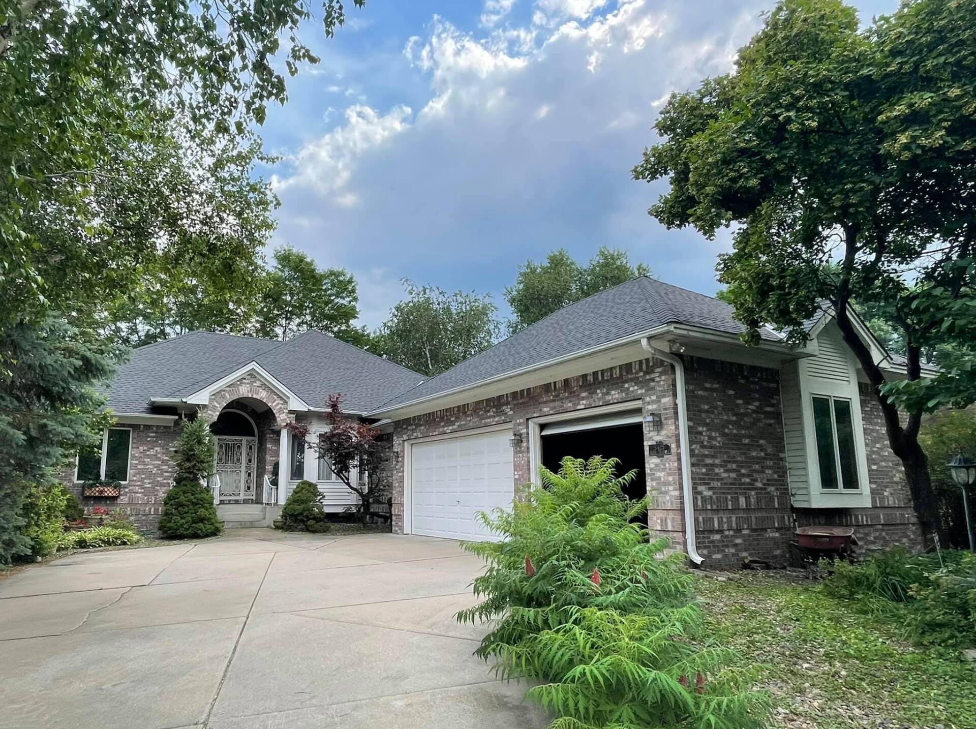 Brick house with a gray roof, garage door open, and a driveway surrounded by trees. Overcast sky.