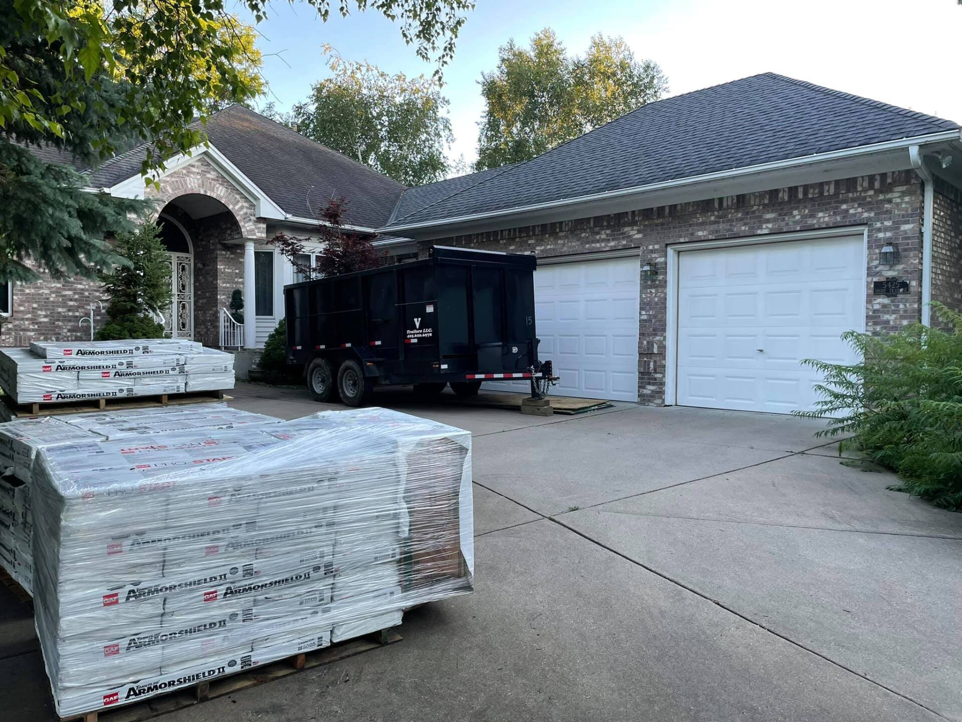 A house with a brick facade and two-car garage, a trailer, and pallets of roofing materials on the driveway.