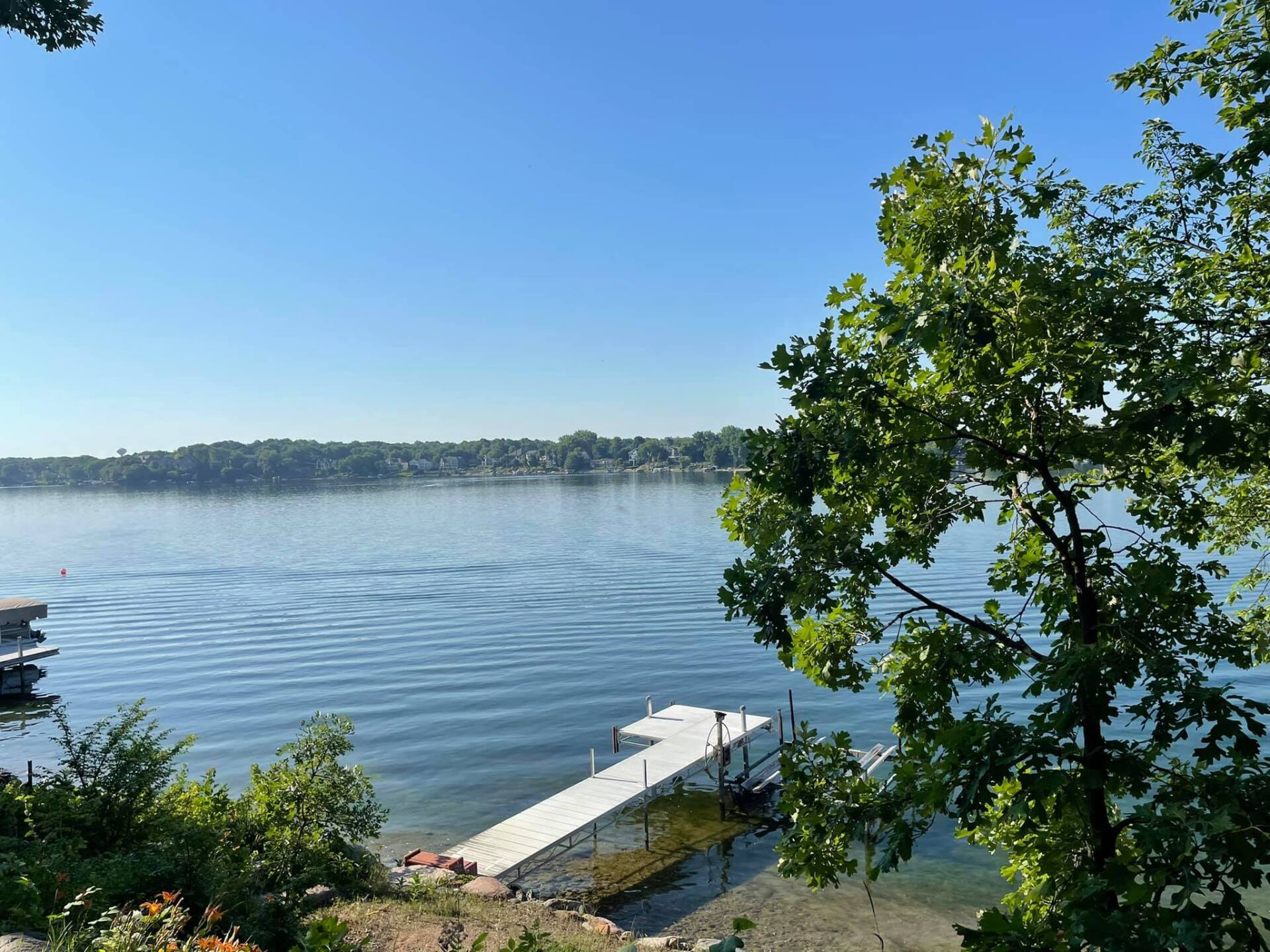 A lake scene with a white dock extending into the water, framed by trees and a clear blue sky.