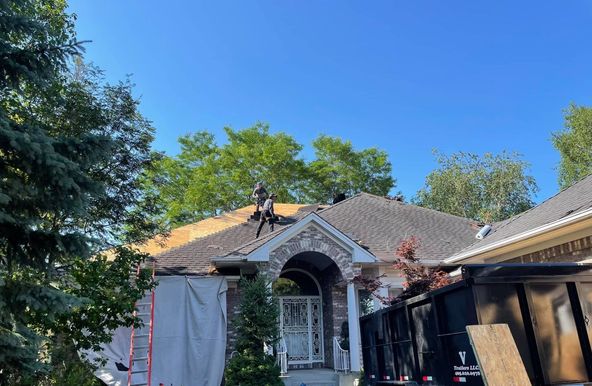 Roof being repaired on a house under a clear blue sky. Workers are on the roof, and a dumpster sits nearby.