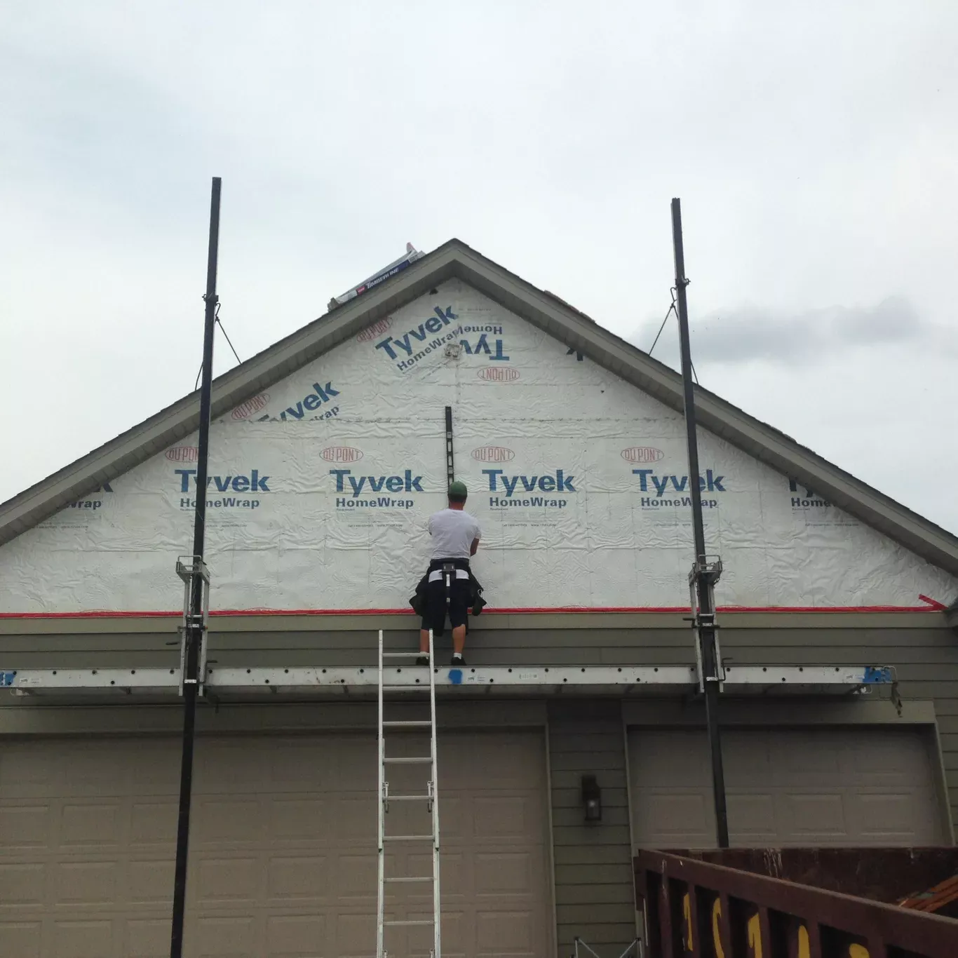 A person on a ladder works on a house's gable end wrapped in Tyvek. Two poles with support beams are set up.