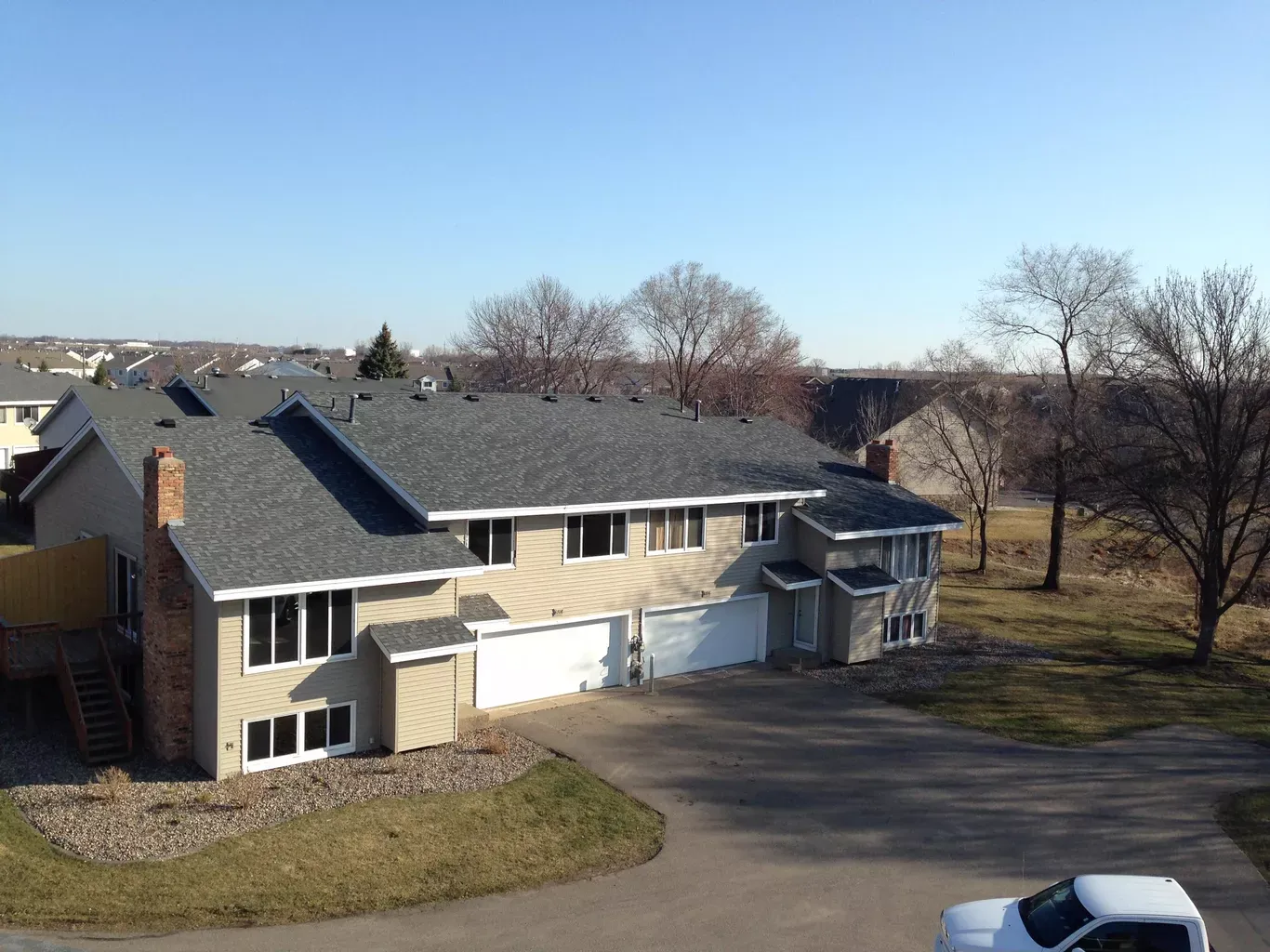 Multi-story beige house with a gray roof, two garage doors, and surrounding brown grass and trees under a blue sky.