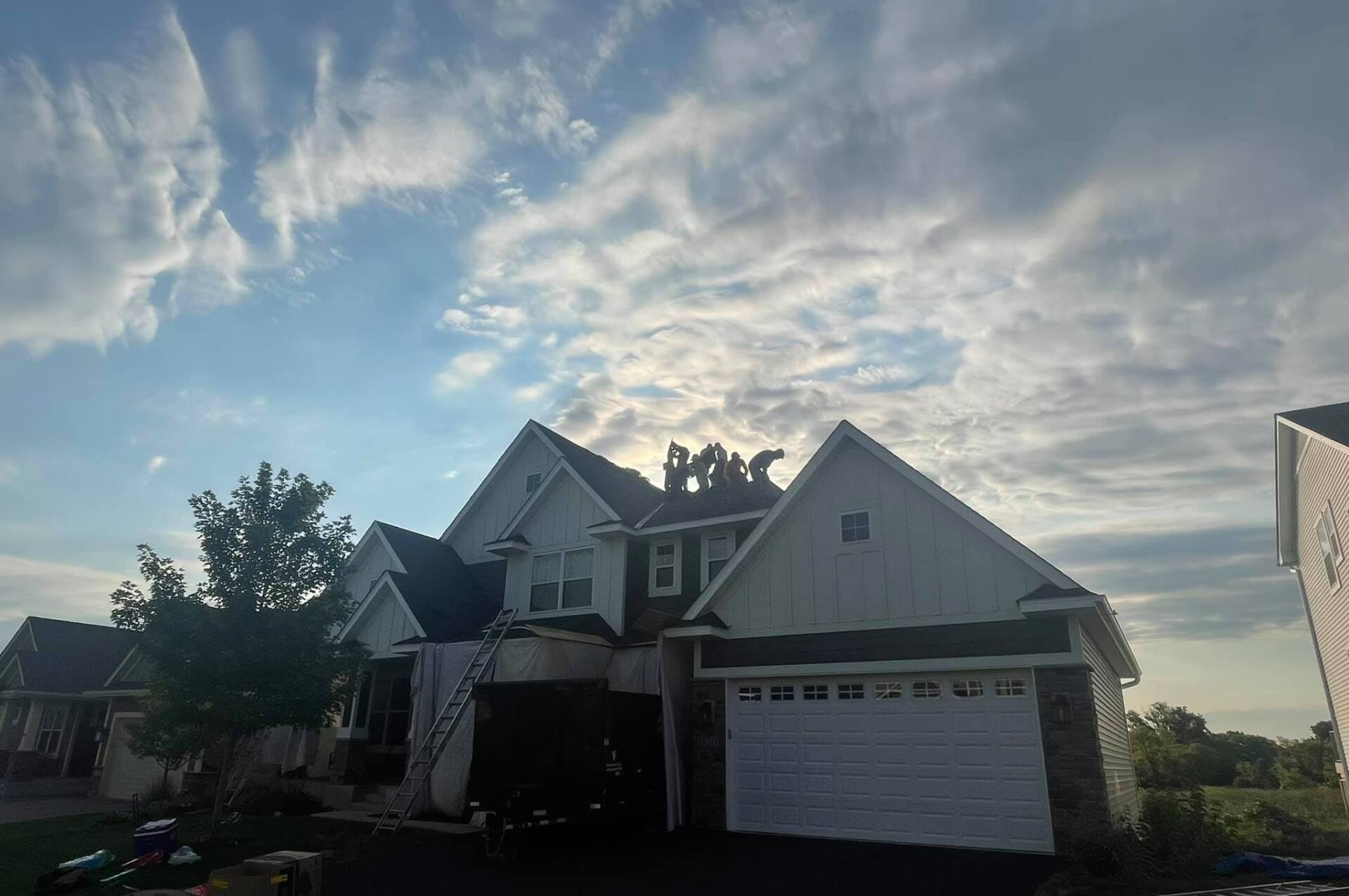 House under construction with workers on the roof; white siding, stone facade, blue sky with clouds.