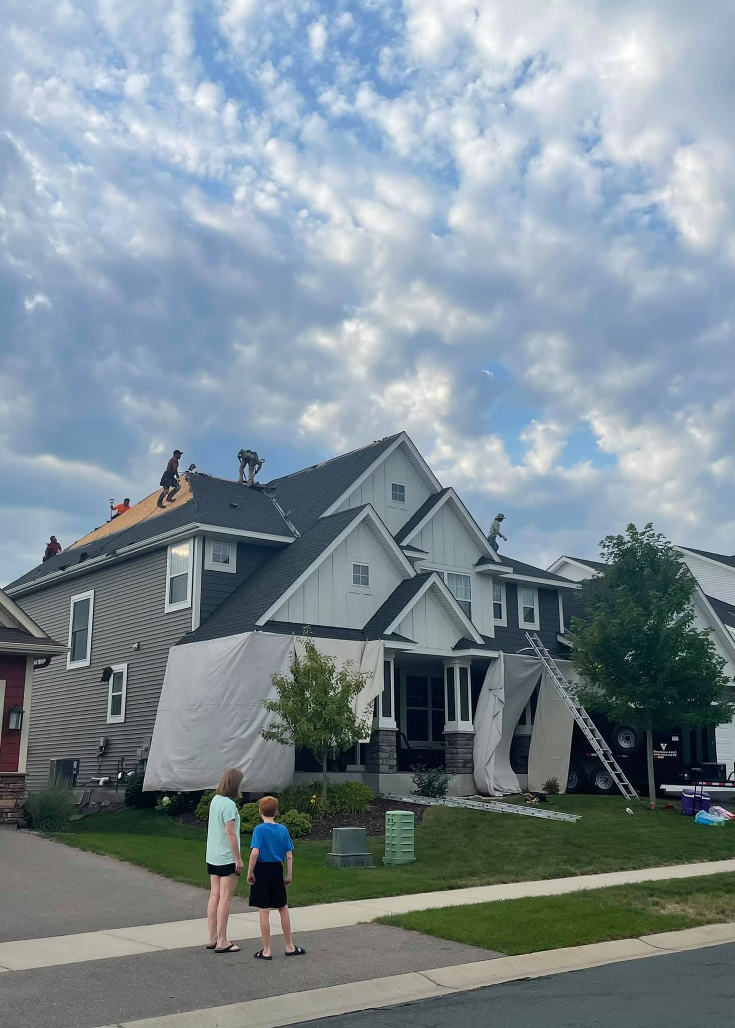 Two children watch as roofers work on a house with protective coverings. The house is gray and white with a dark roof under a cloudy sky.