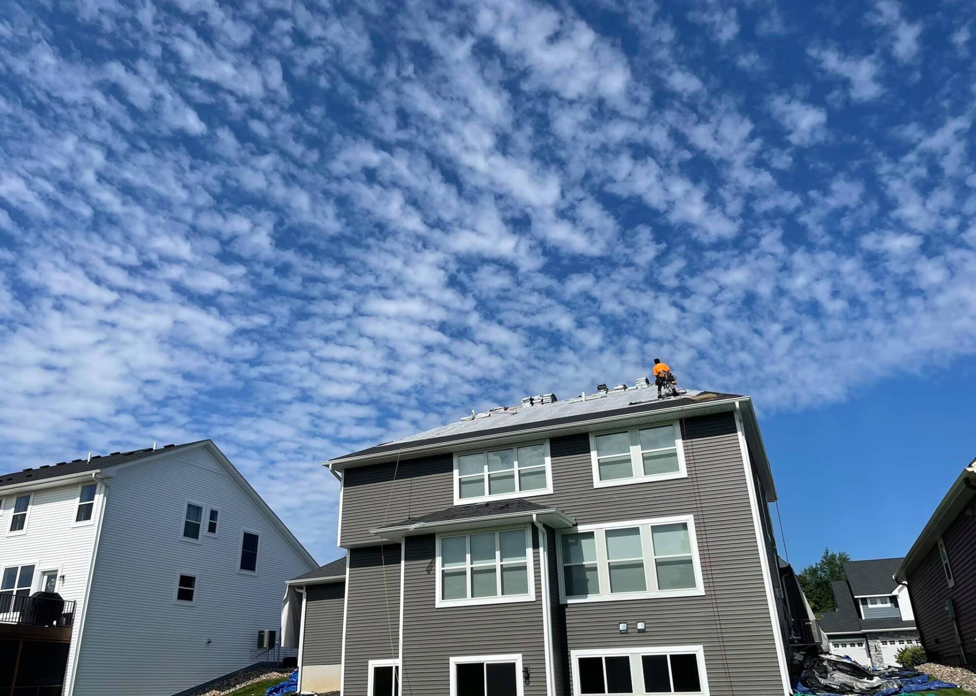 Two-story gray house with white trim under a blue sky with puffy clouds; a person works on the roof.