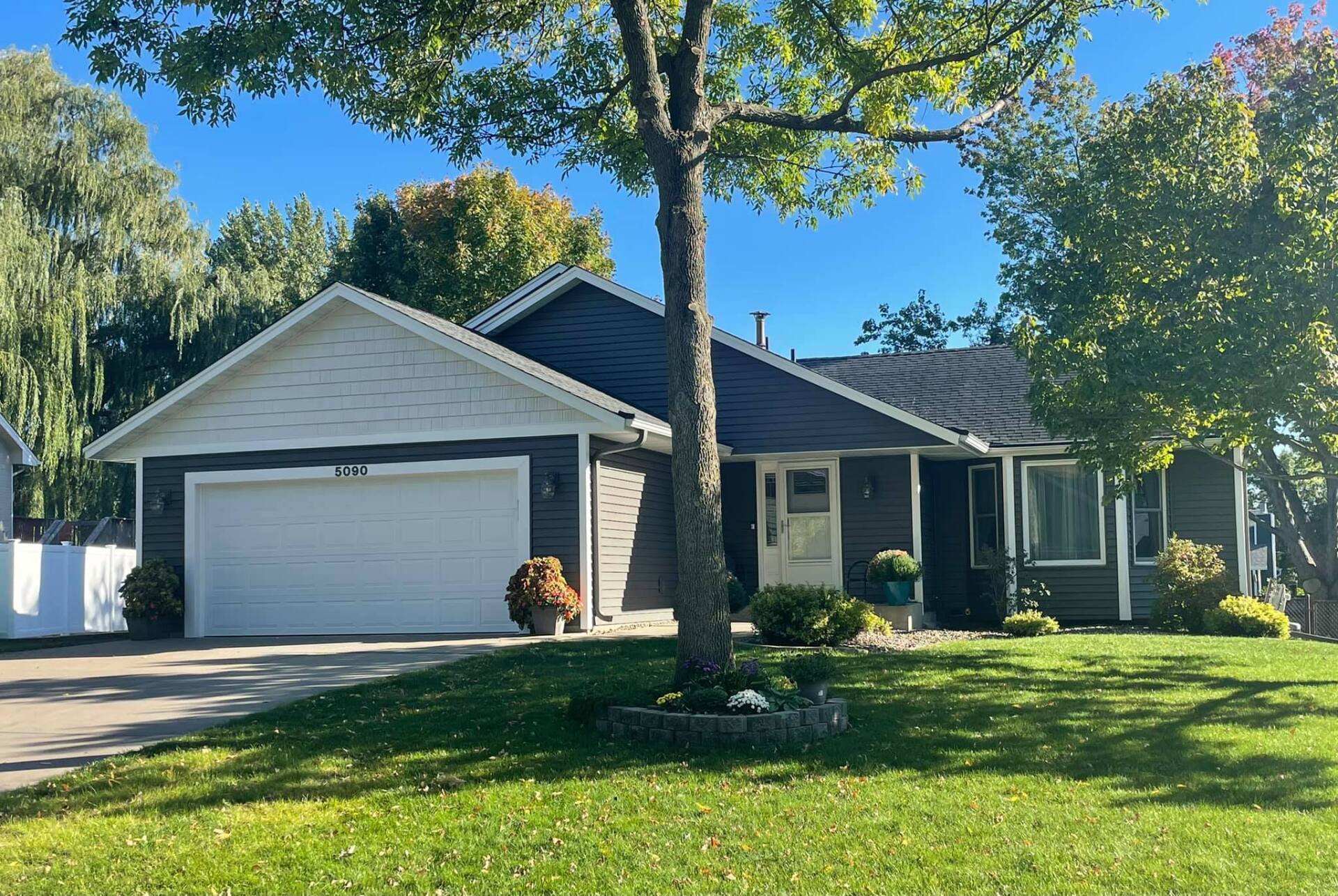 A single-story house with dark gray siding and a white garage door. A tree is in front of the house, and there is green grass.