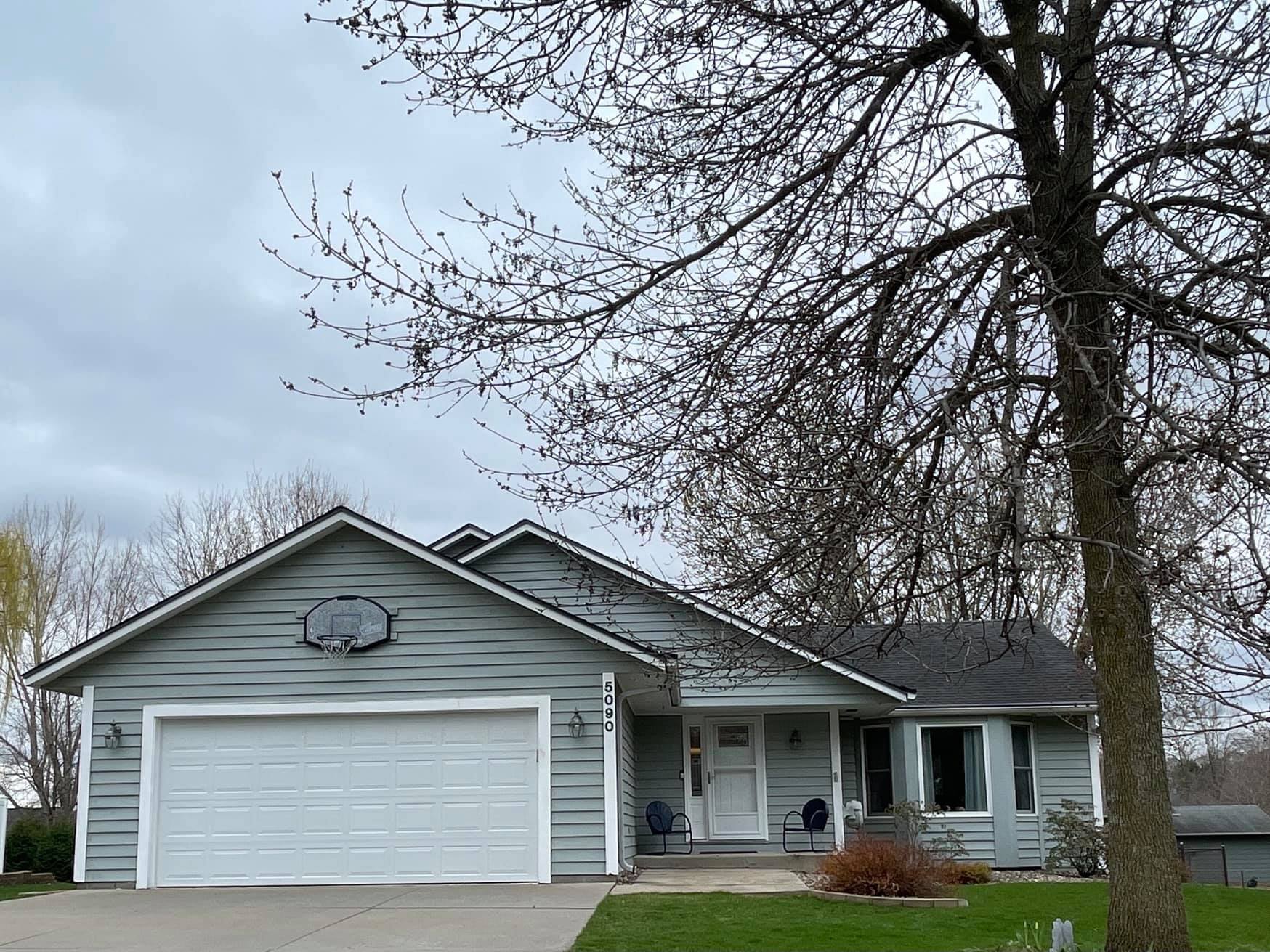 A gray house with a basketball hoop over the garage door, under a cloudy sky. A bare tree is in the foreground.
