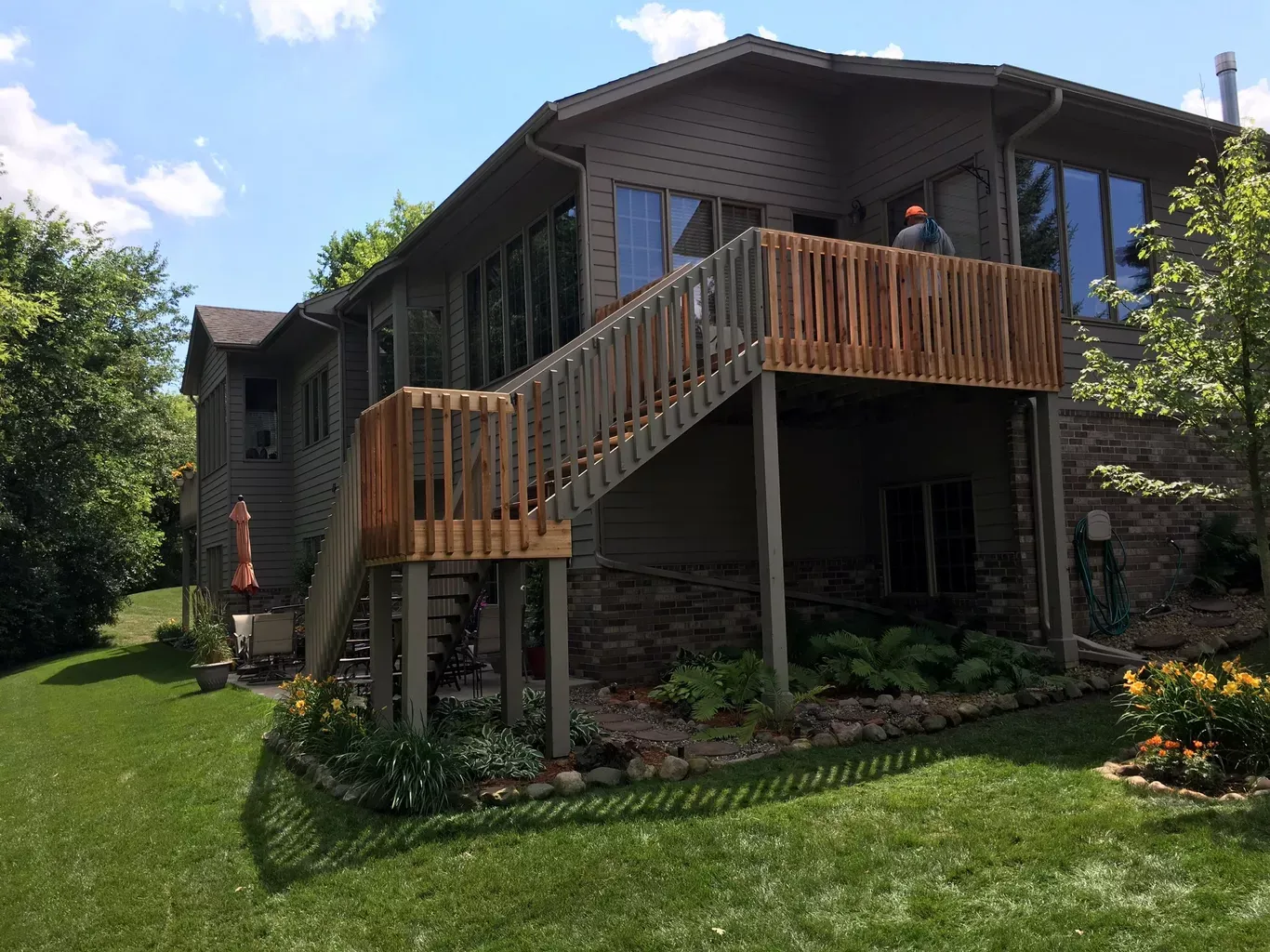 House with a wooden deck and stairs, surrounded by green lawn and trees. The house has large windows and a stone facade.