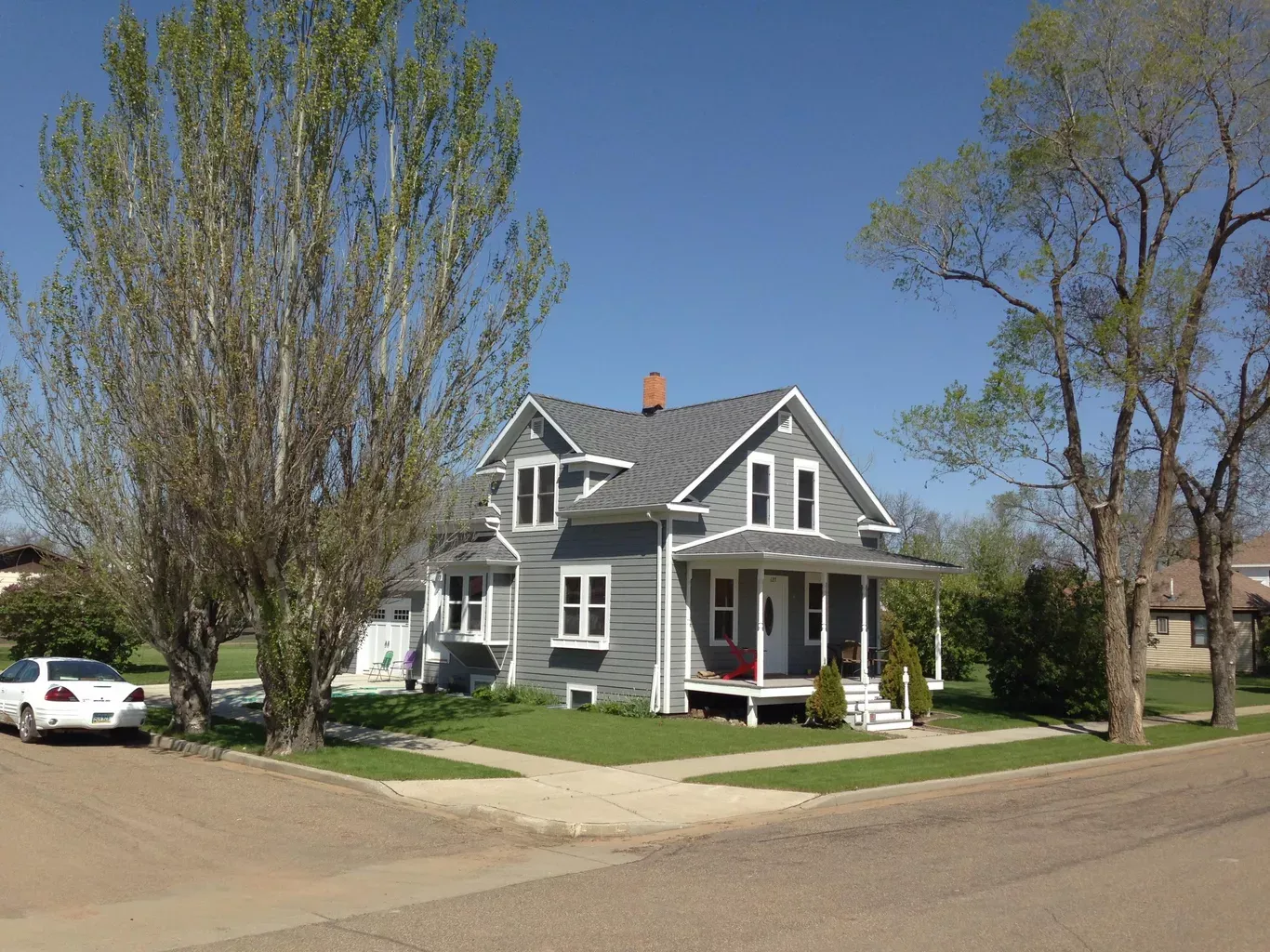 Gray two-story house on a corner lot, with a white porch, and trees on a sunny day. A car is parked on the street.