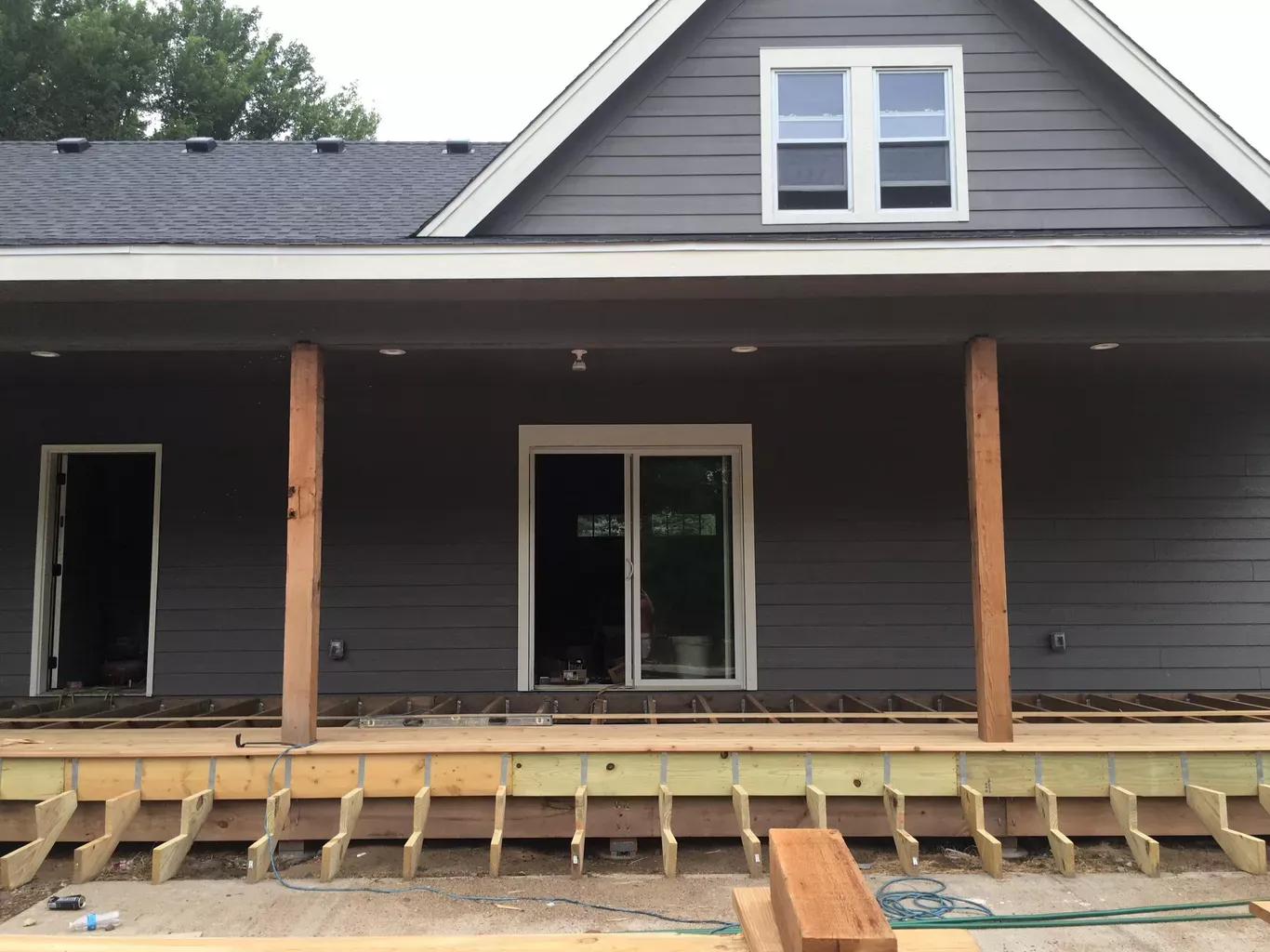 House exterior under construction. Grey siding, unfinished wooden porch frame. Two wooden posts support the roof overhang.