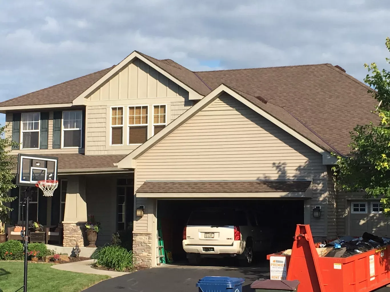 A two-story house with a car in the garage, a basketball hoop, and a construction dumpster. The house has brown roofing and tan siding.