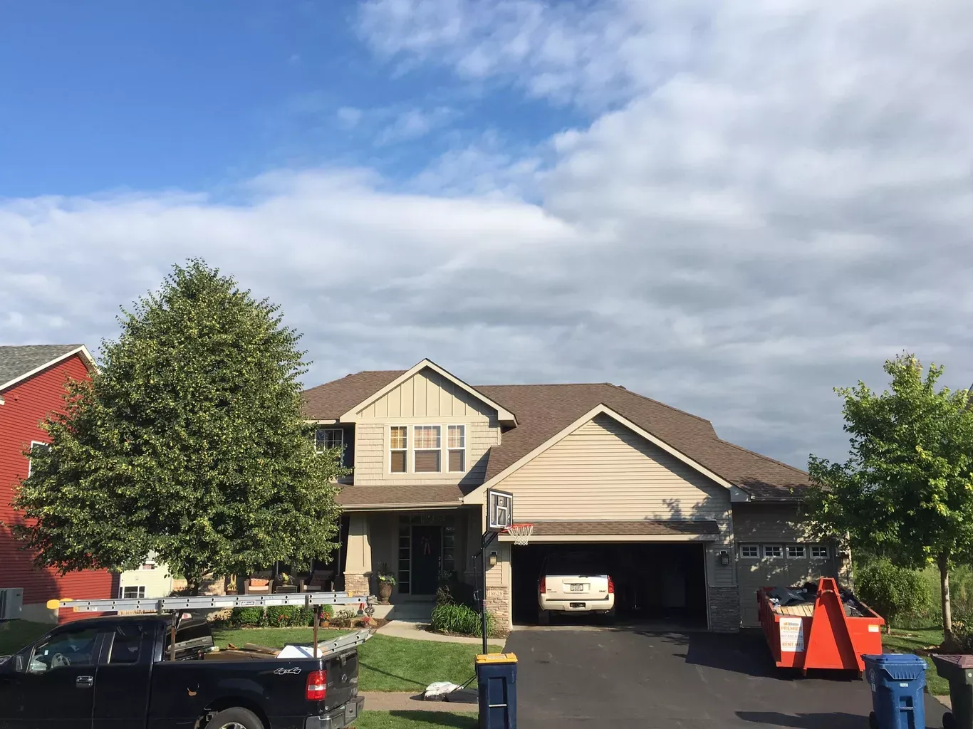 Two-story house with beige siding and a brown roof; a car is parked inside the open garage. A tree and a black truck are in the foreground.