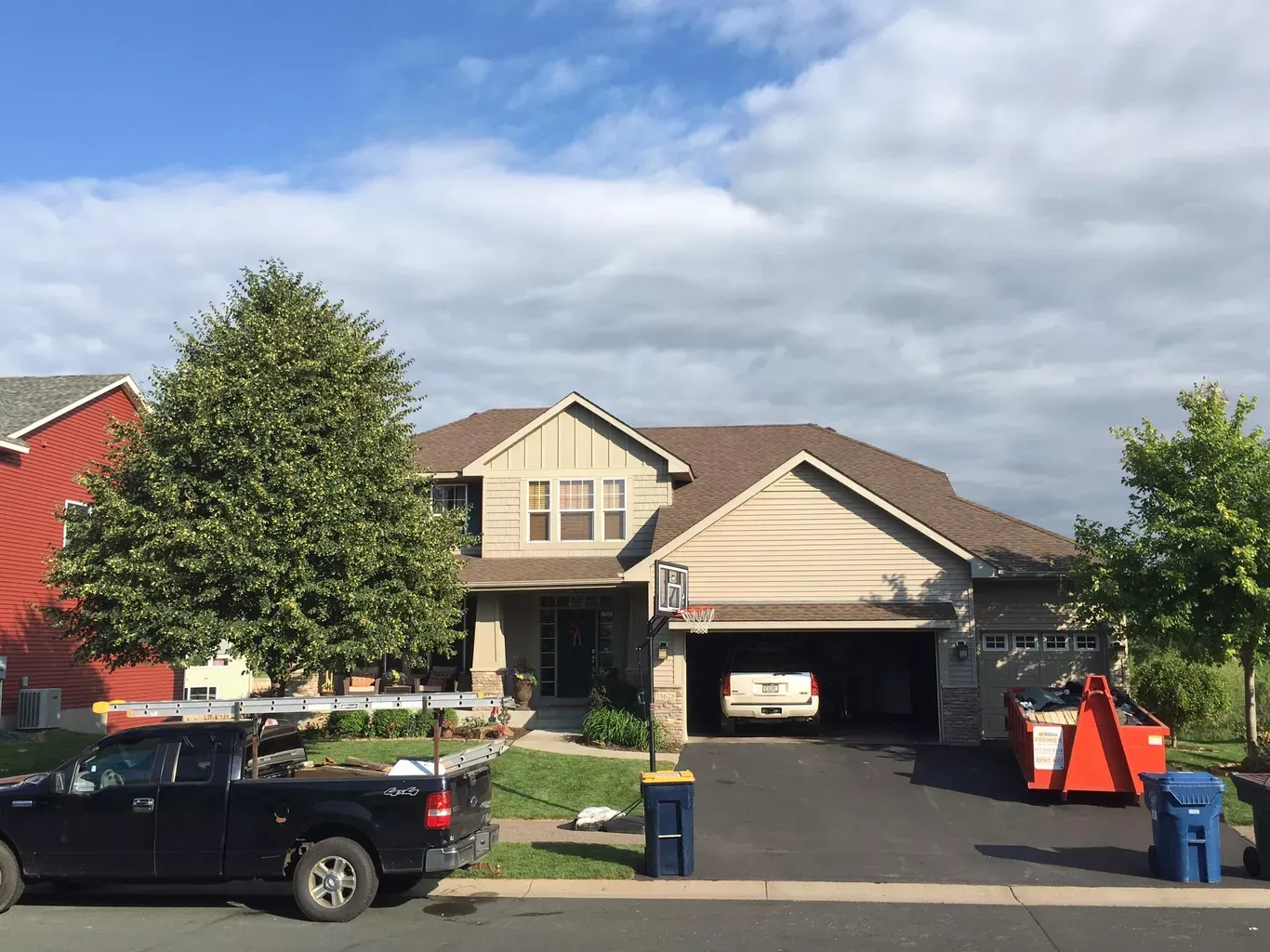 Two-story house with tan siding and a brown roof, a car in the garage, a tree, and a pickup truck parked on the street.