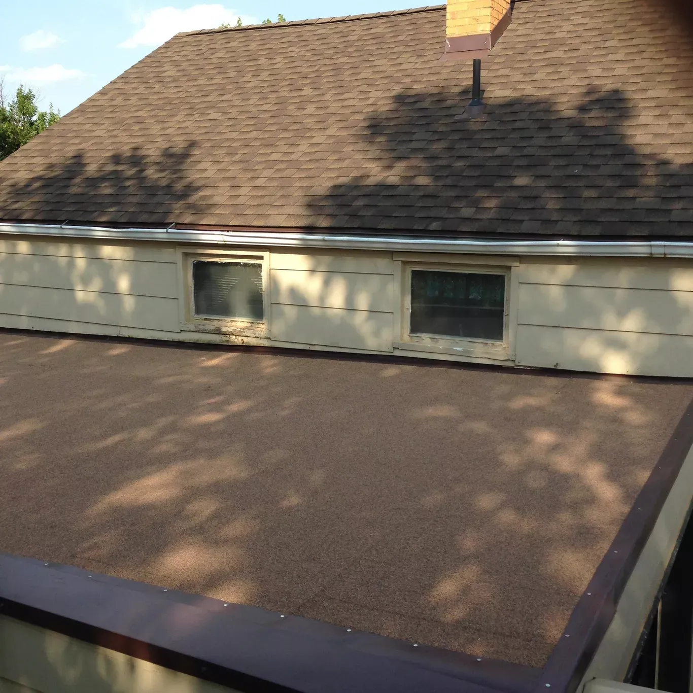 A brown, flat roof with a granular surface, bordered by a dark metal edge, and a brown shingled roof in the background.