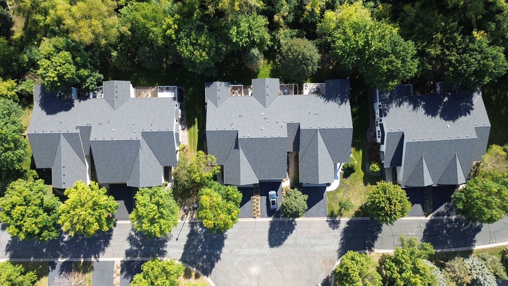 Aerial view of three townhouses with gray roofs, a road, and surrounding green trees.