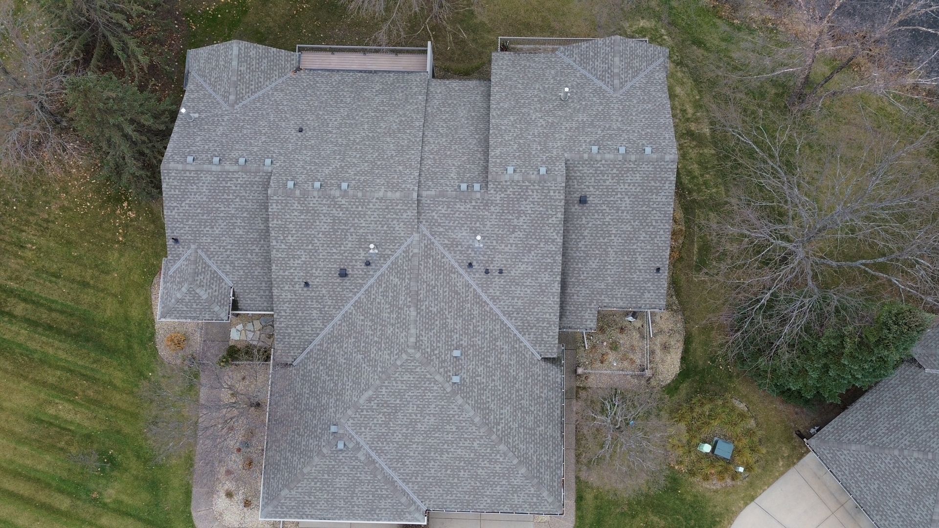 Overhead view of a gray shingled roof with several sections, trees, and green grass.