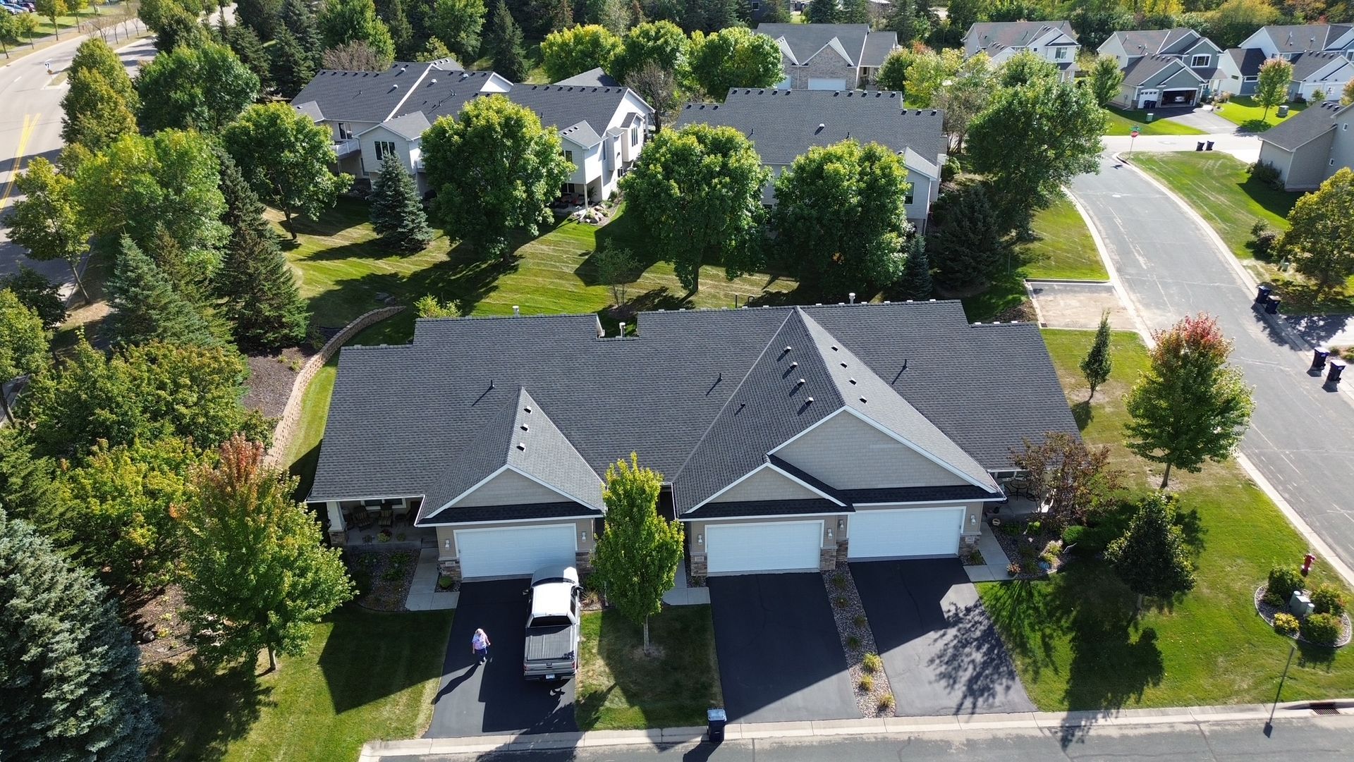 Aerial view of a duplex with black roofs and attached garages. Trees and a curved road surround the homes in a suburban setting.