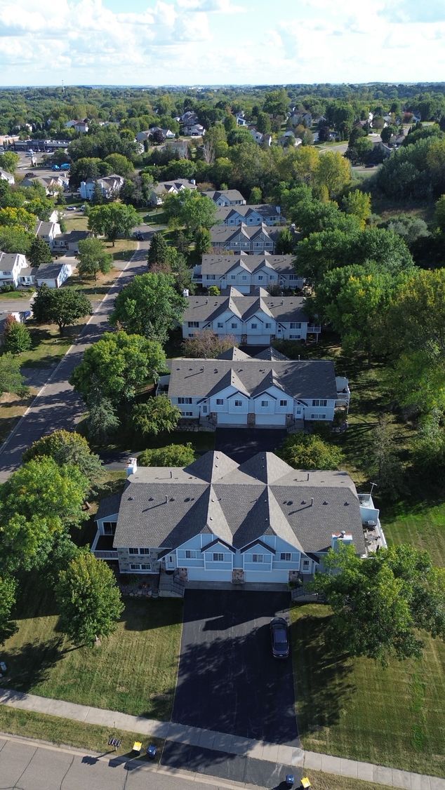 Row of light-colored townhouses with gray roofs, nestled among green trees, set in a suburban area on a sunny day.