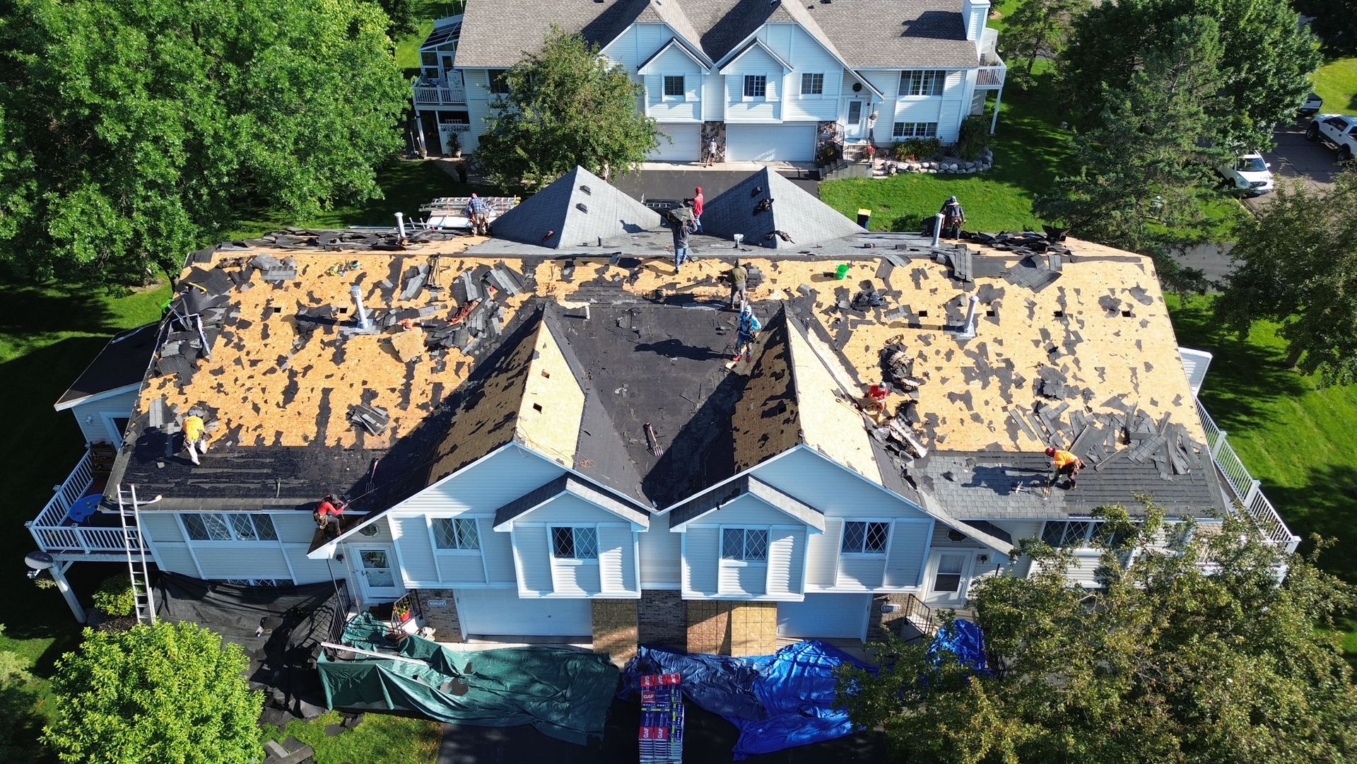 Workers replacing shingles on a large, light blue house with a complex roof structure; green lawn surrounds.