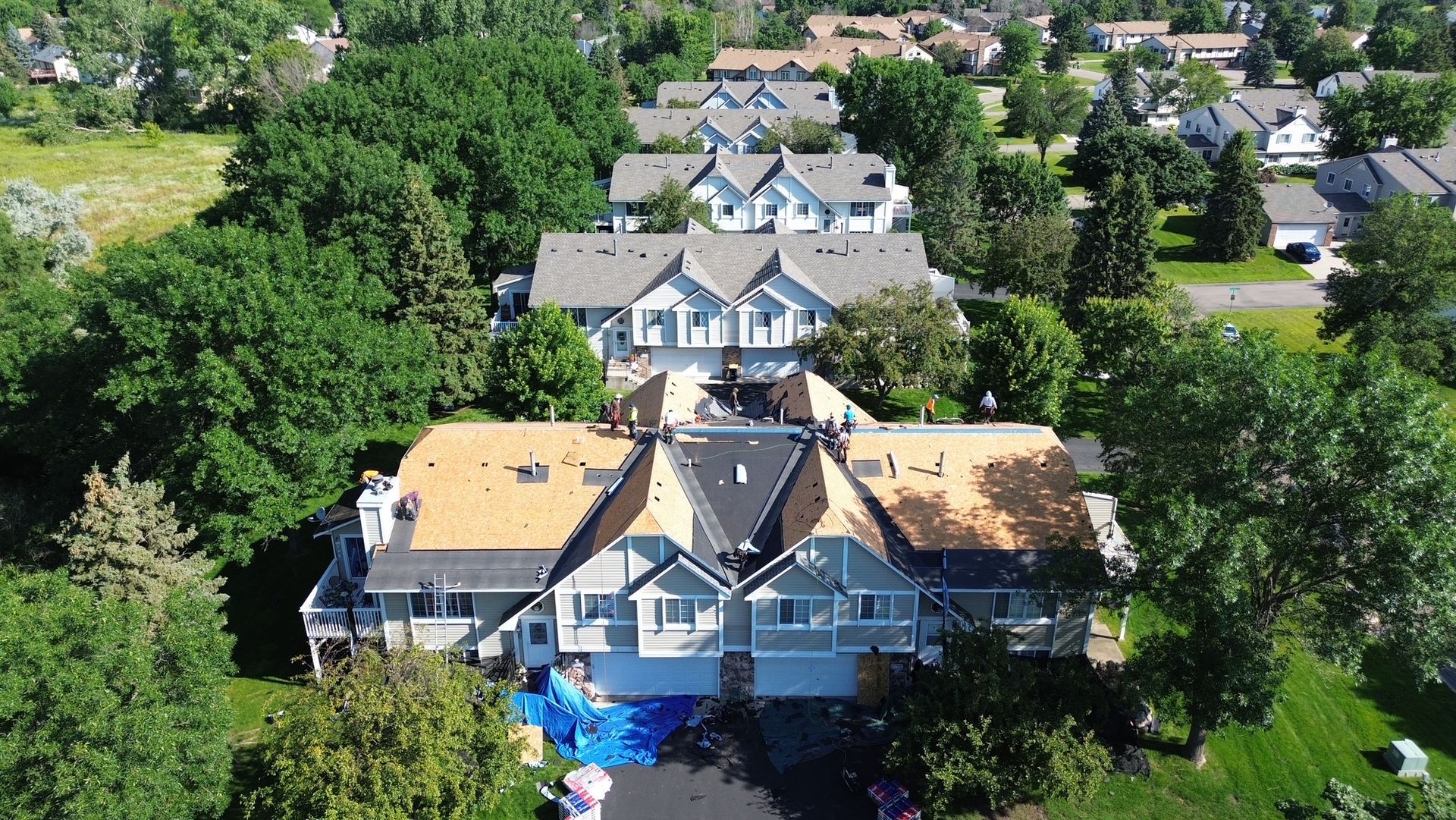 Aerial view of townhouses; roofers are working on a roof with missing shingles. Blue tarps cover the ground.