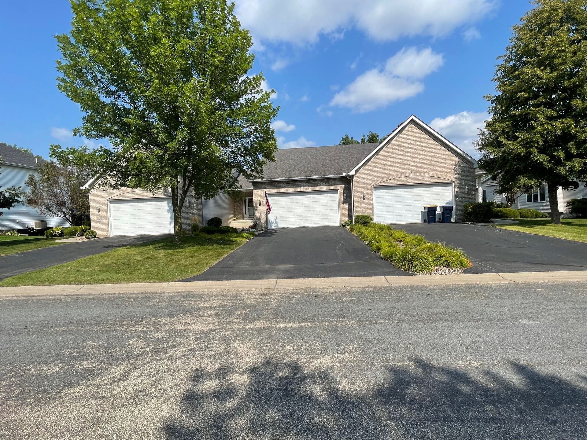 Three-car garage attached to a brick house, with black asphalt driveways, green grass, and a cloudy blue sky.