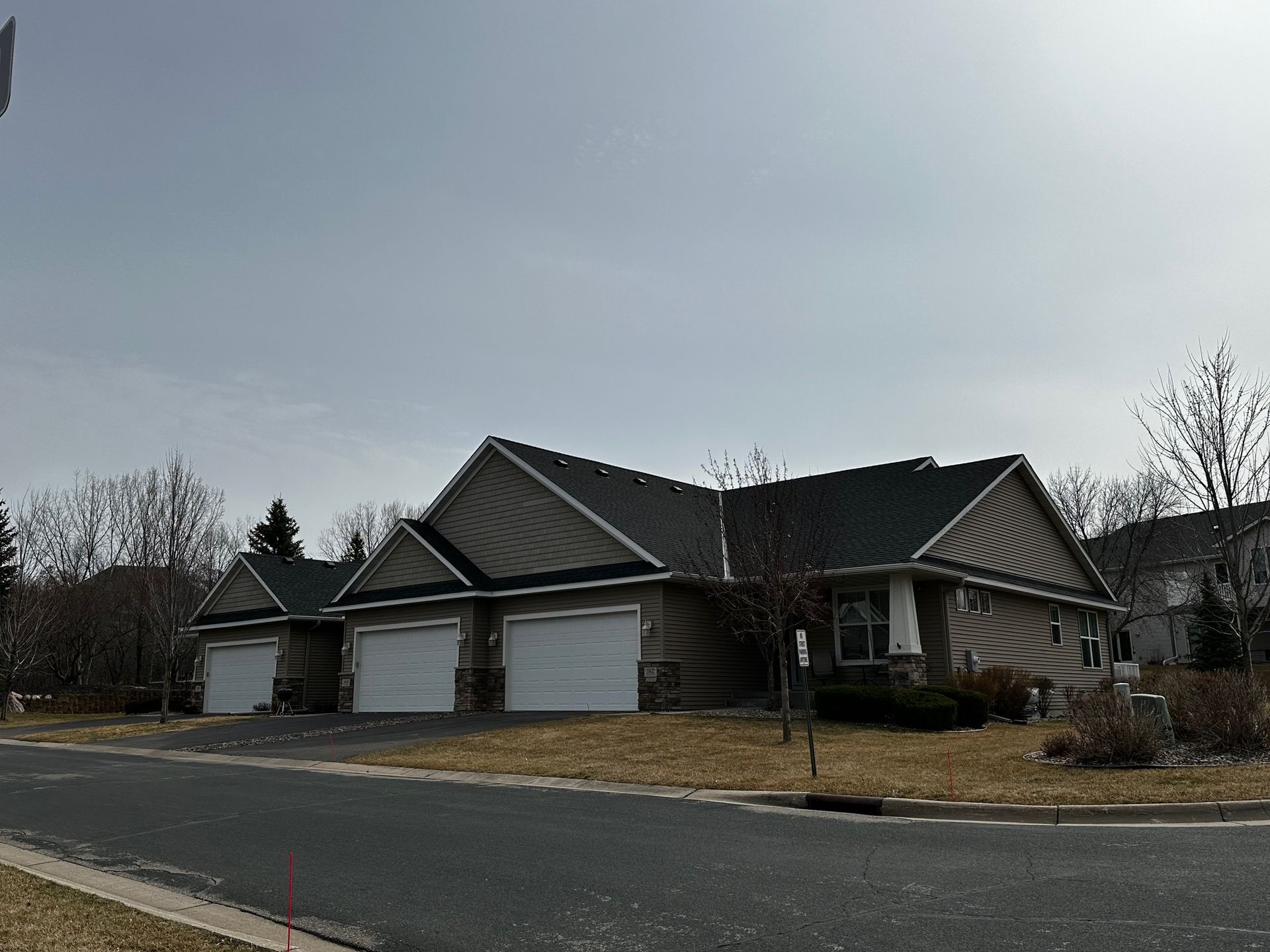 A one-story house with a three-car garage on a street with bare trees and a gray sky. The house is brown with a dark roof.