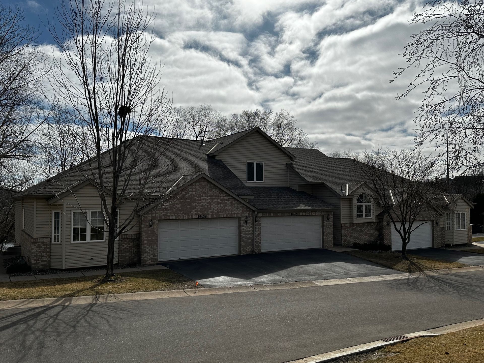 A multi-unit home with three garage doors and a brick and beige exterior under a cloudy sky. Leafless trees are in the foreground.