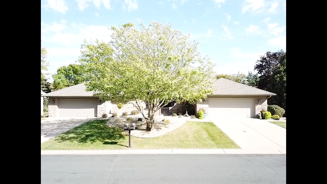 A split-level, beige house with two garage doors, a large tree in front, and a green lawn under a blue sky.