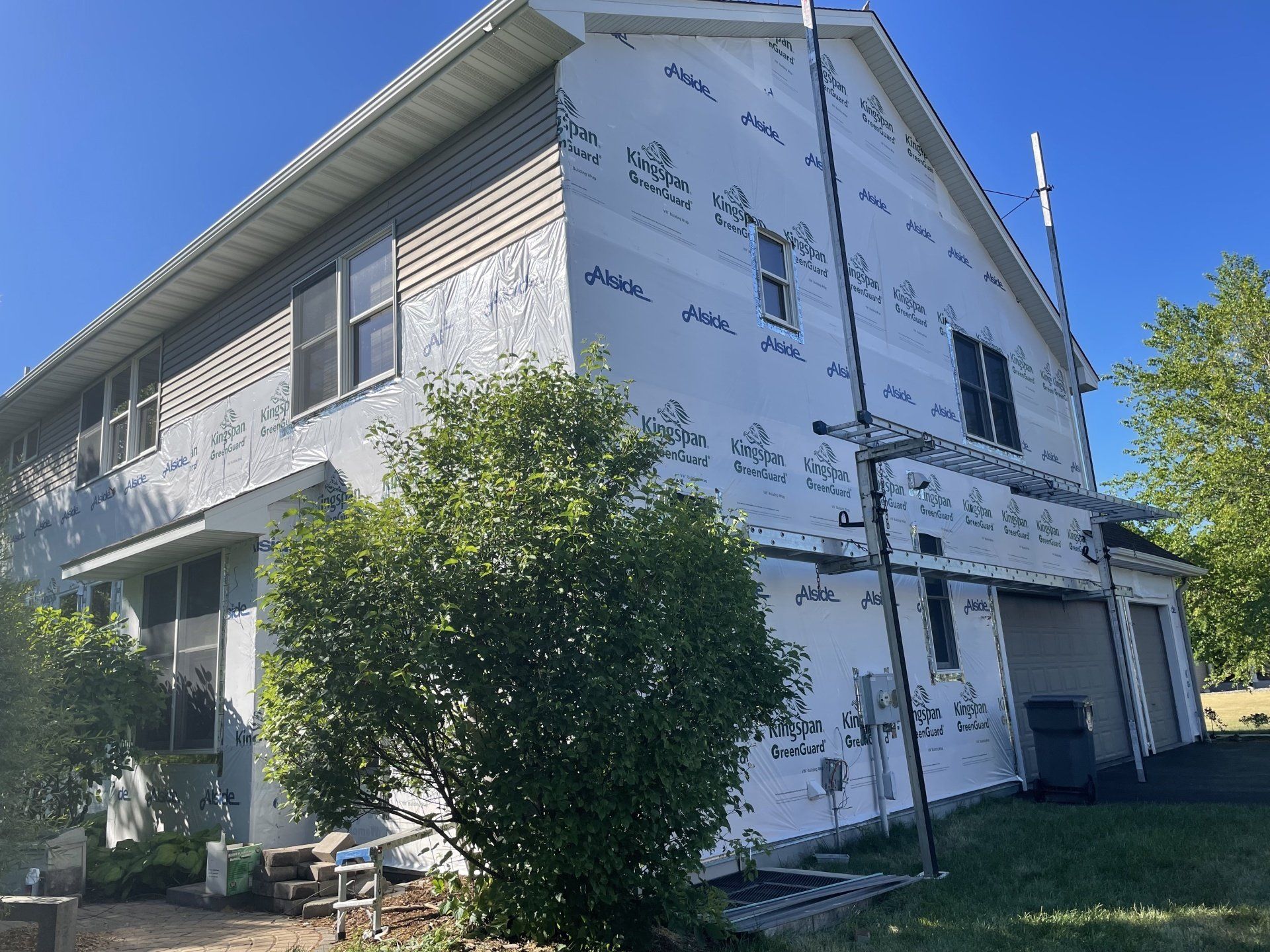 House exterior partially covered with white protective wrap; blue sky overhead. A ladder leans against the side.