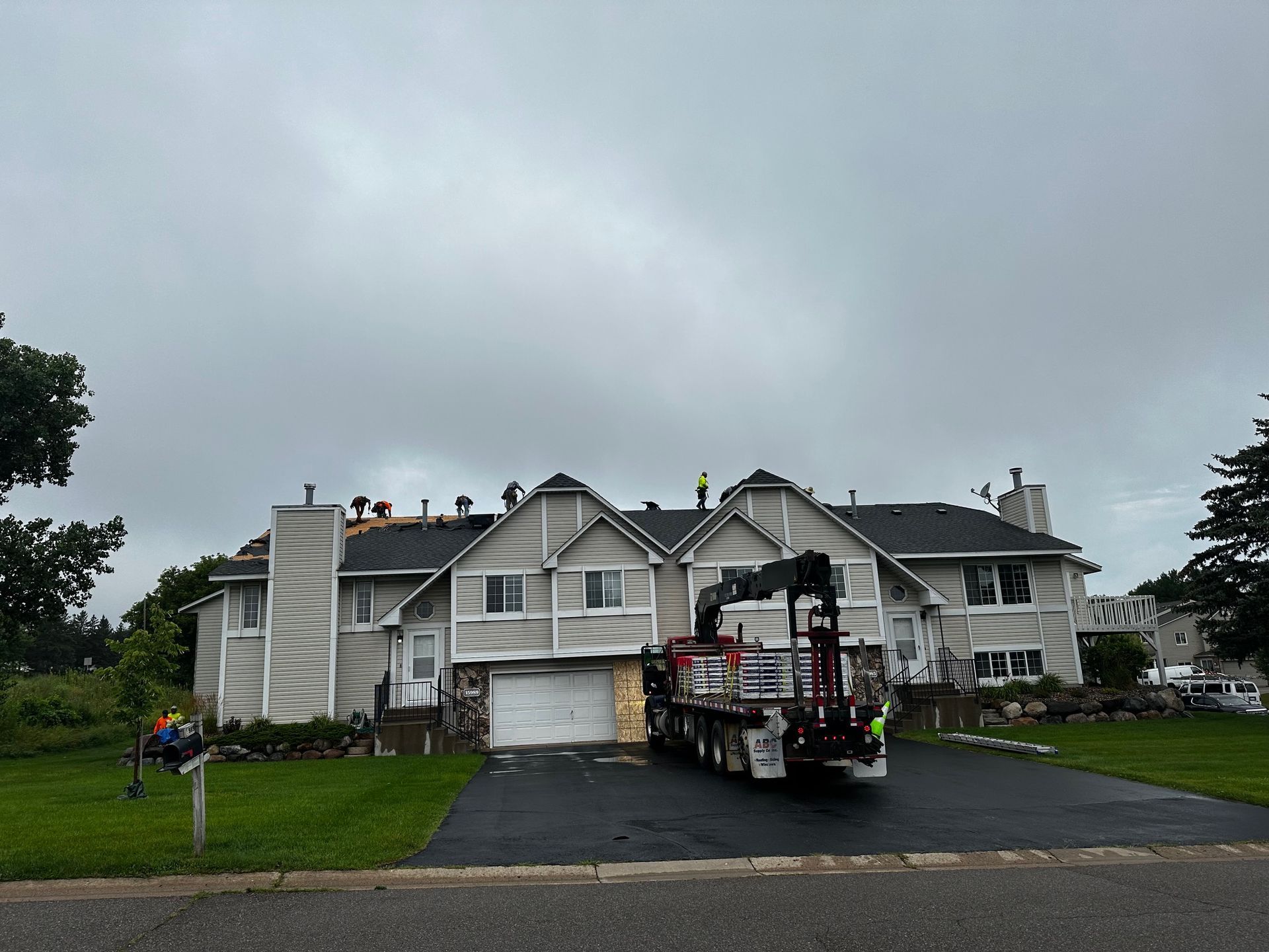 A crane truck in front of a house under construction. The roof has exposed framing, and workers are likely present. Overcast sky.