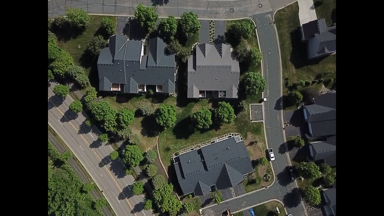 Overhead view of a residential area with dark-roofed houses, roads, and green trees casting shadows on the grass.