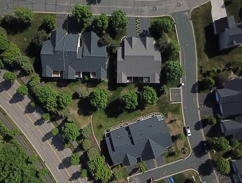 Aerial view of residential homes with dark roofs, surrounded by trees and roads.