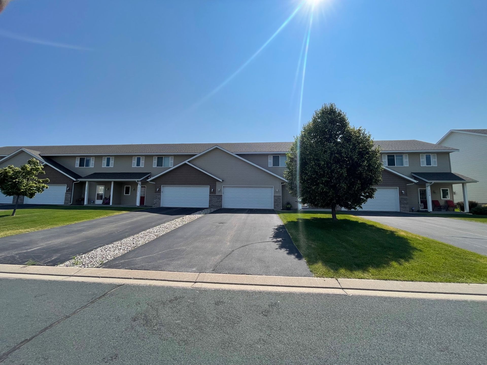 Row of townhouses with attached garages on a sunny day.  Driveways lead to the garages, with a grassy area and tree in front.