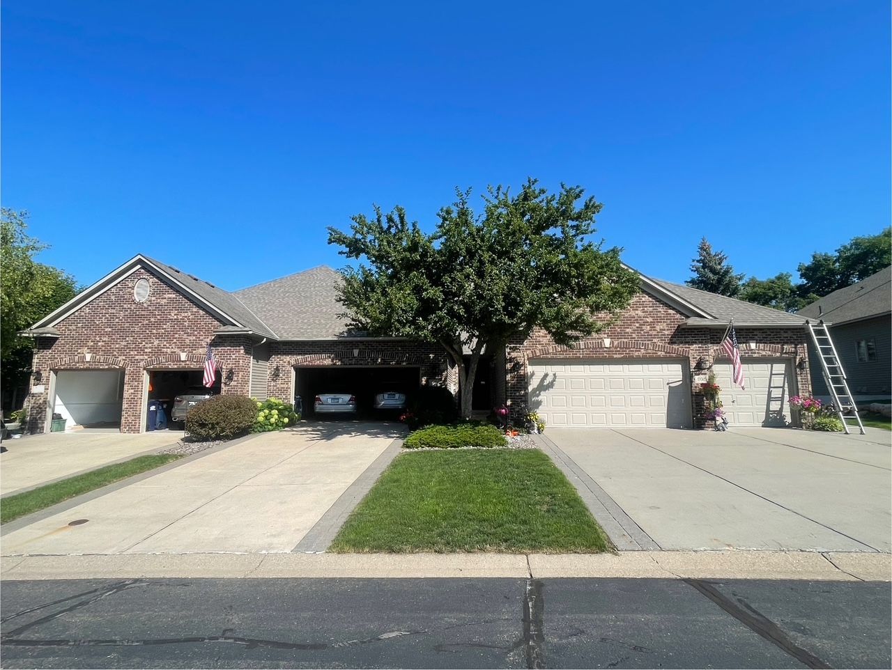 Two brick homes with attached garages under a clear blue sky. A tree and green lawn sit between driveways.