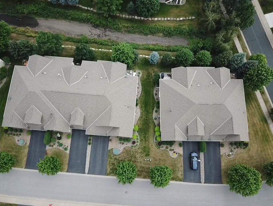 Aerial view of two adjacent houses with dark driveways, tan roofs, and surrounding trees and bushes. A black car is parked in one driveway.