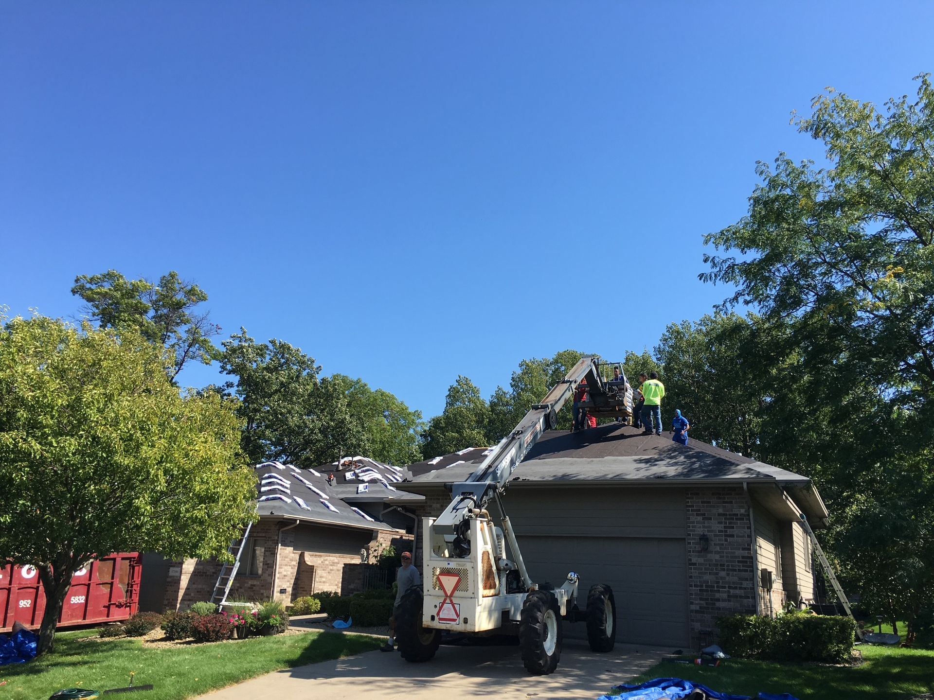 Roofers on a house roof with equipment; clear blue sky and green trees in the background.