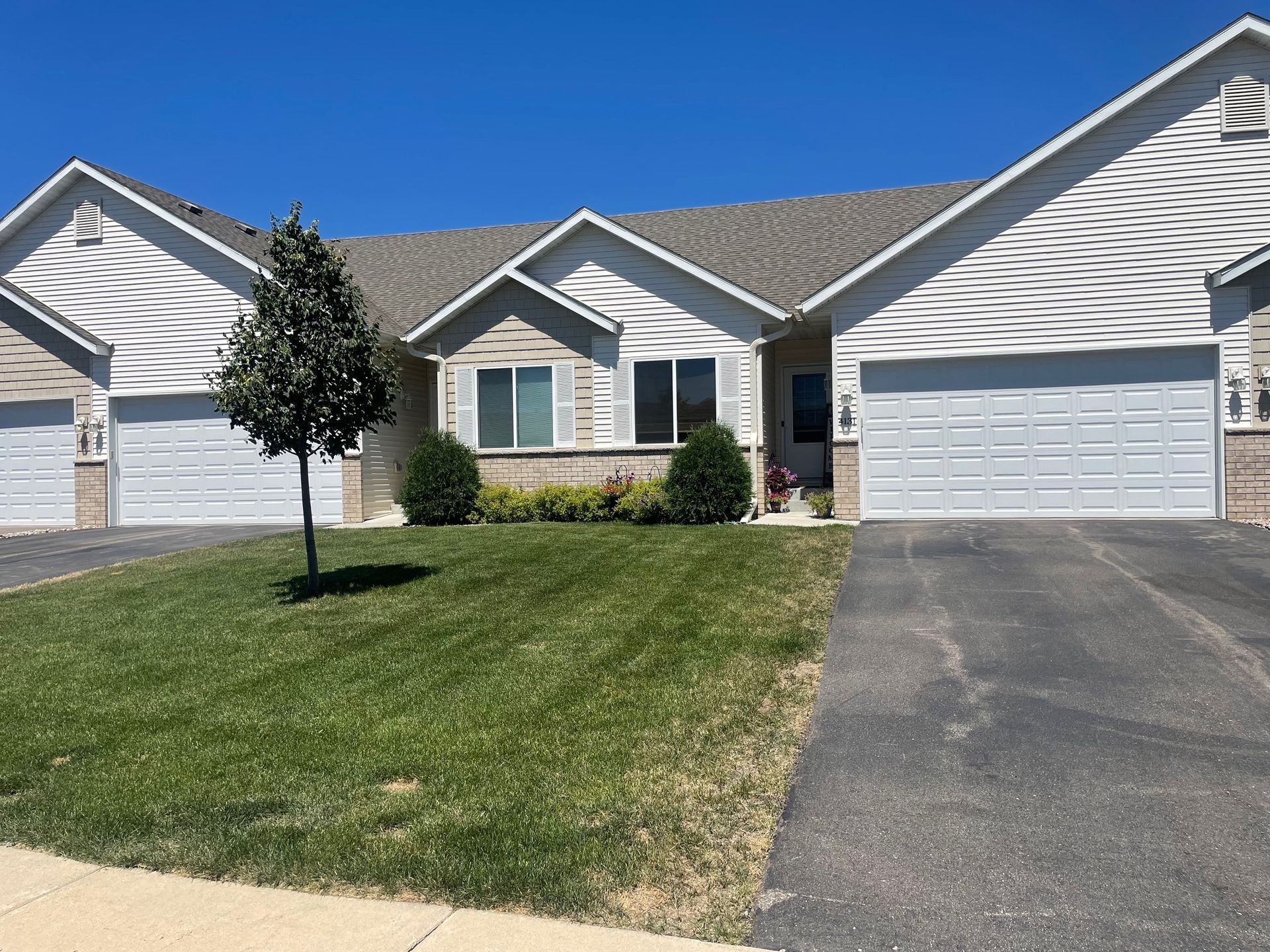 Row of attached houses with white siding, gray garages, and a small tree in front on a sunny day.