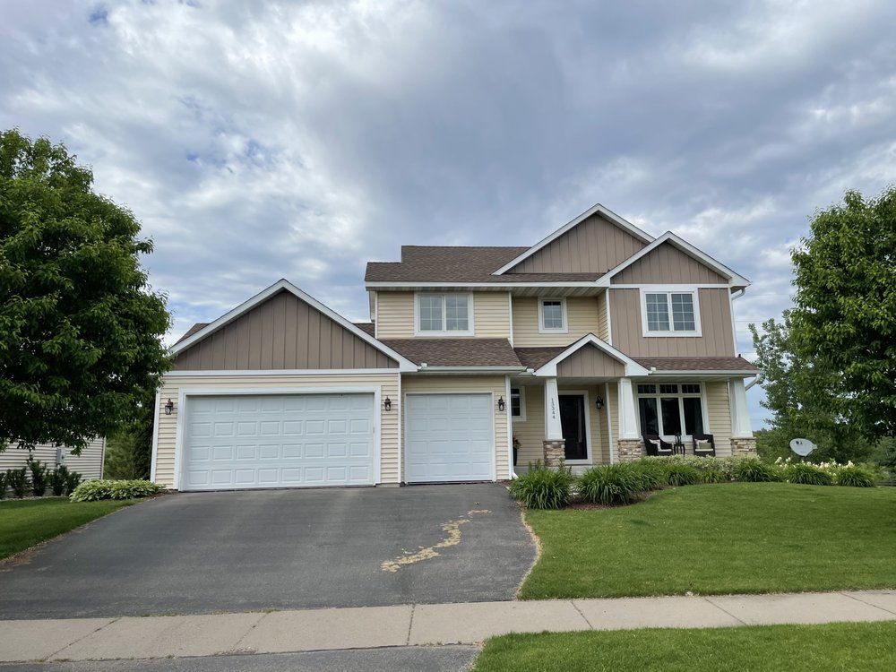Two-story house with a brown roof, tan siding, and two garage doors. A driveway and green lawn lead to the house under a cloudy sky.