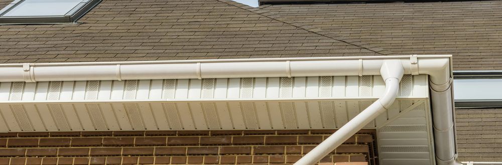 White gutters on a house with brown roof shingles and a brick facade. A downspout is visible on the right.