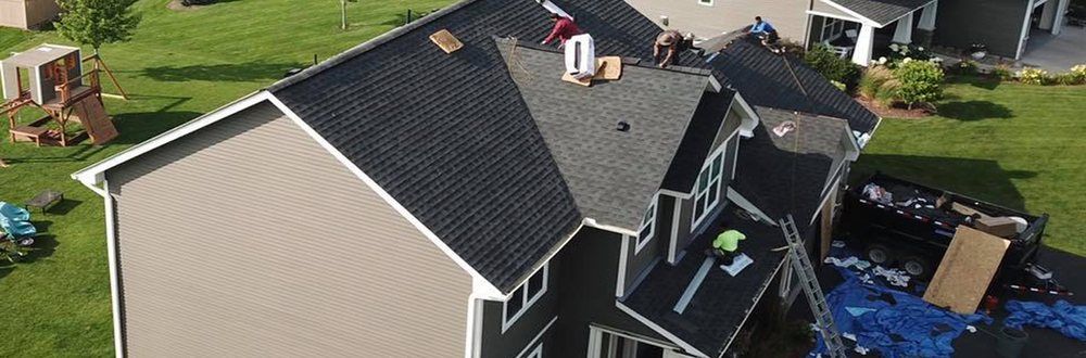 Aerial view of a house with workers on the roof. The house has a dark gray roof with tan siding, surrounded by green grass.