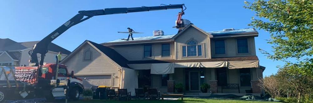 A crane is lifting roofing materials to a two-story house, presumably for roof repair or replacement. Workers are on the roof.