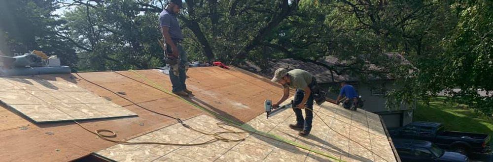 Two roofers install plywood sheeting on a roof. One is hammering, and the other stands nearby. Green trees are in the background.