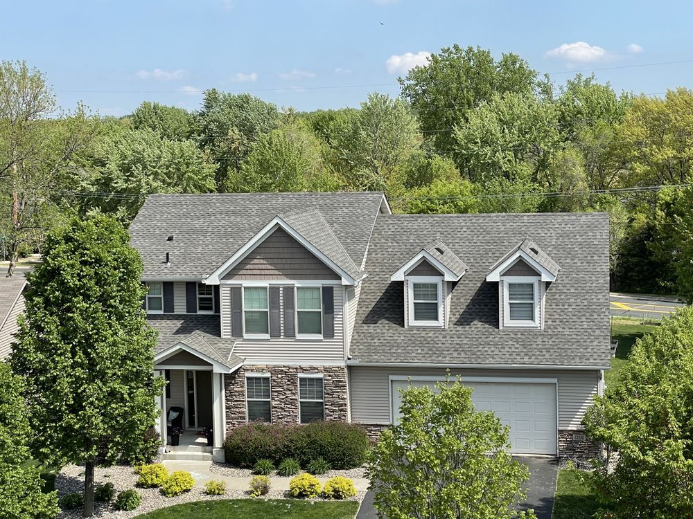 Two-story house with gray roof, stone and stucco facade, green trees in the background, sunny day.