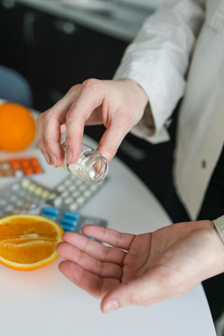 Person pouring pills from a bottle into their hand, near medicine and orange slices.