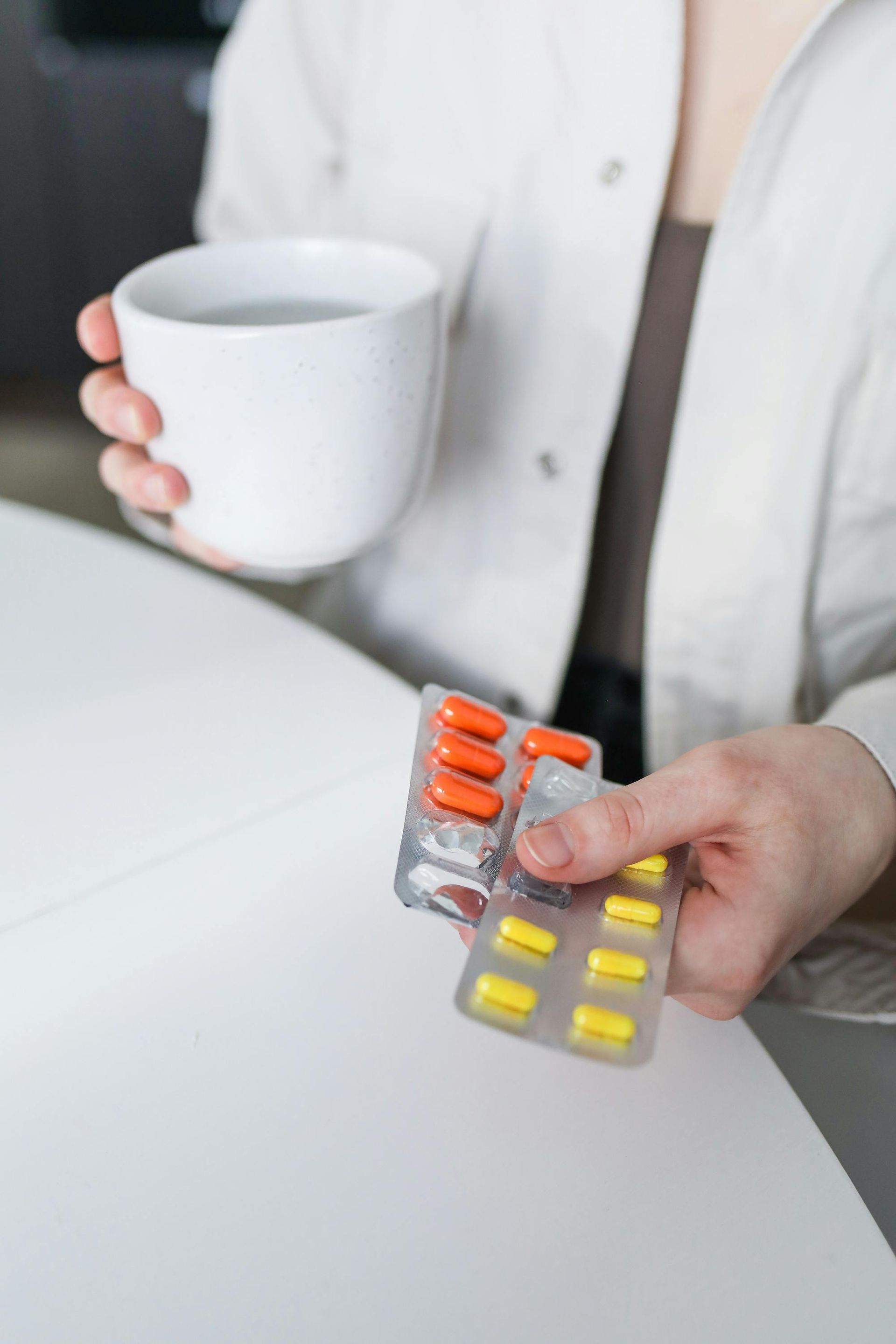 Person holding pills in one hand, cup in the other. Orange and yellow capsules in blister packs.