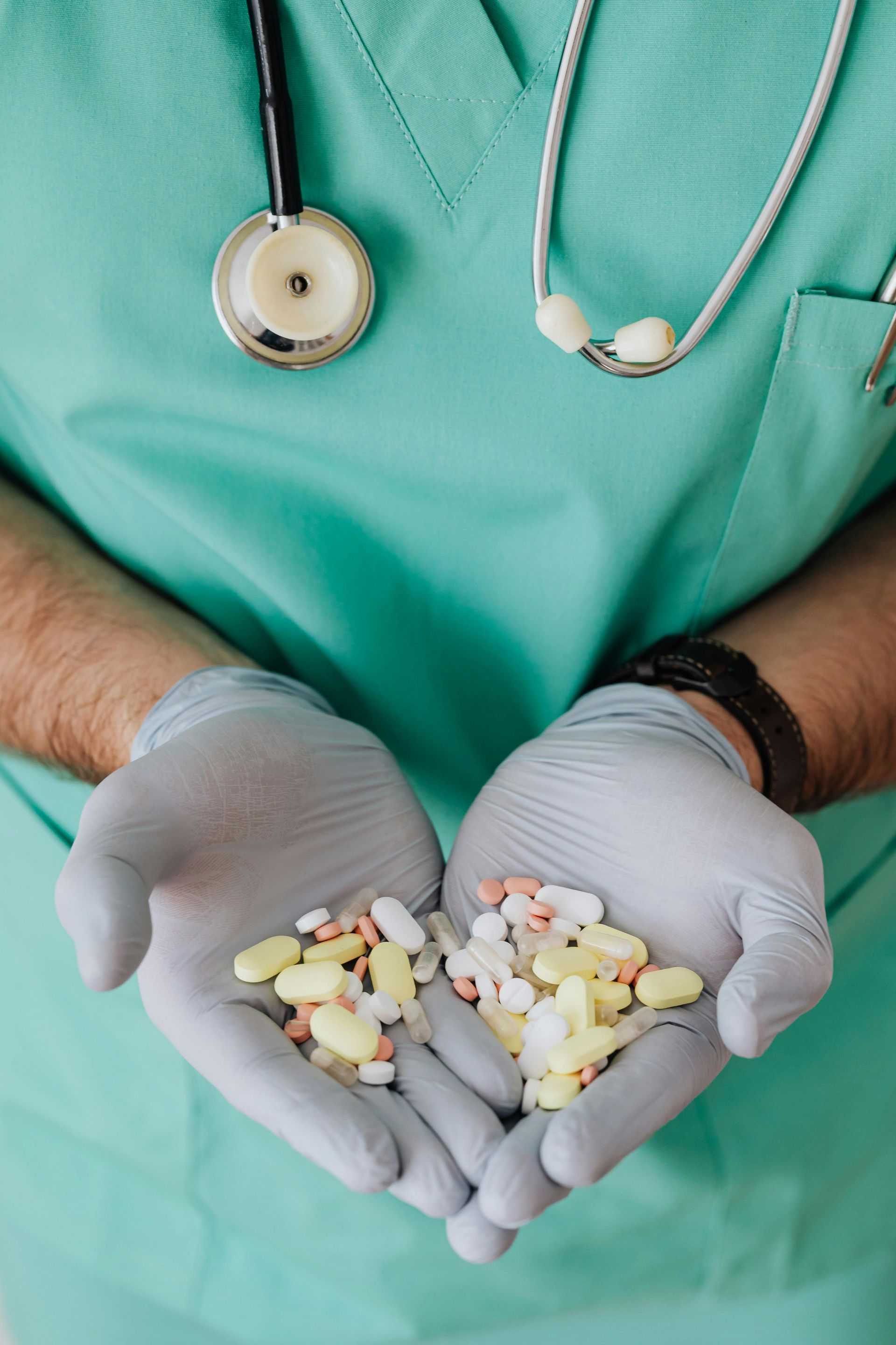 Doctor holding pills in gloved hands, stethoscope around the neck, wearing a green scrub.