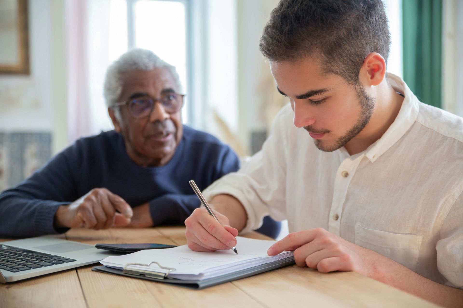 Young man writing on a clipboard while an older man looks on; both are seated at a table.